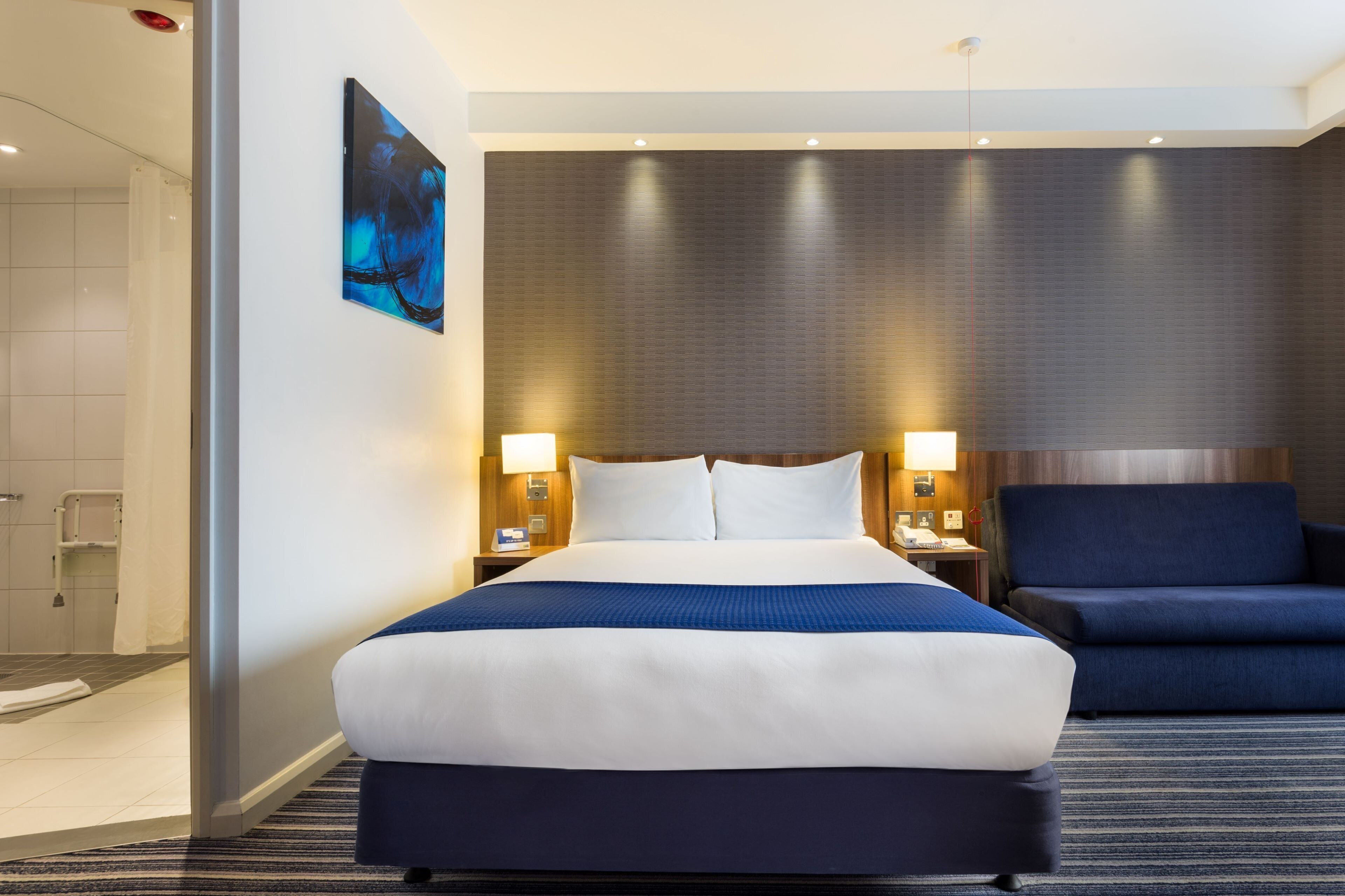 Hotellihuone, jossa on suuri sänky, kaksi yöpöytää, tuoli ja suihkutila.