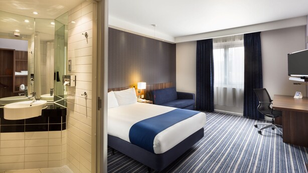 A hotel room with a bed, a desk, a chair, a TV, a bathroom with a sink and mirror, and a window with curtains. - Holiday Inn Express Colchester by IHG (Colchester)