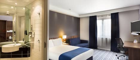 A hotel room with a bed, a desk, a chair, a TV, a bathroom with a sink and mirror, and a window with curtains.