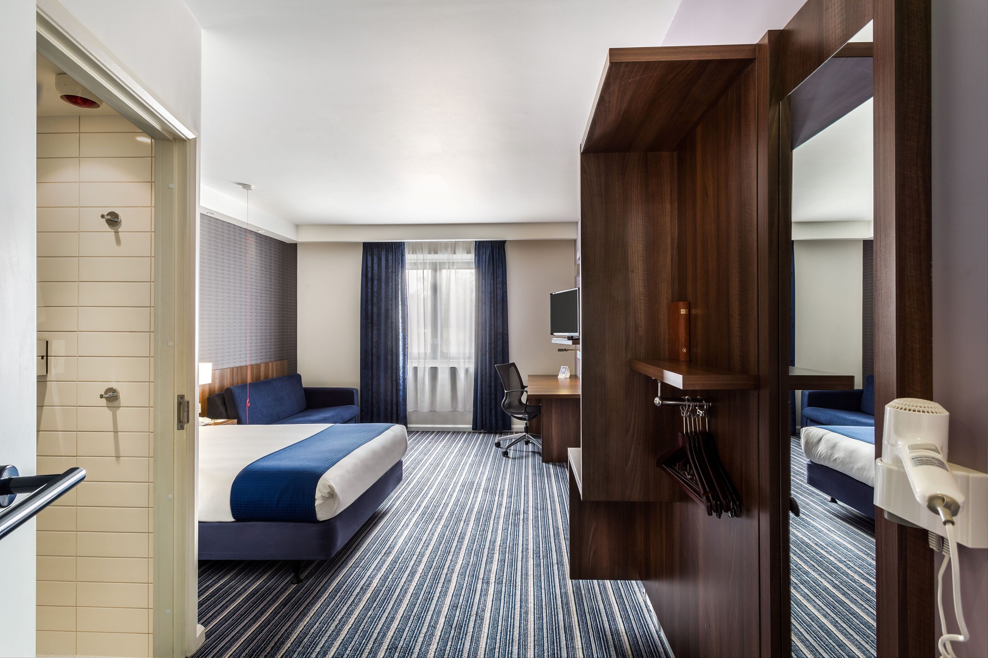 Una habitación de hotel con cama, escritorio, silla, espejo y zona de ducha.