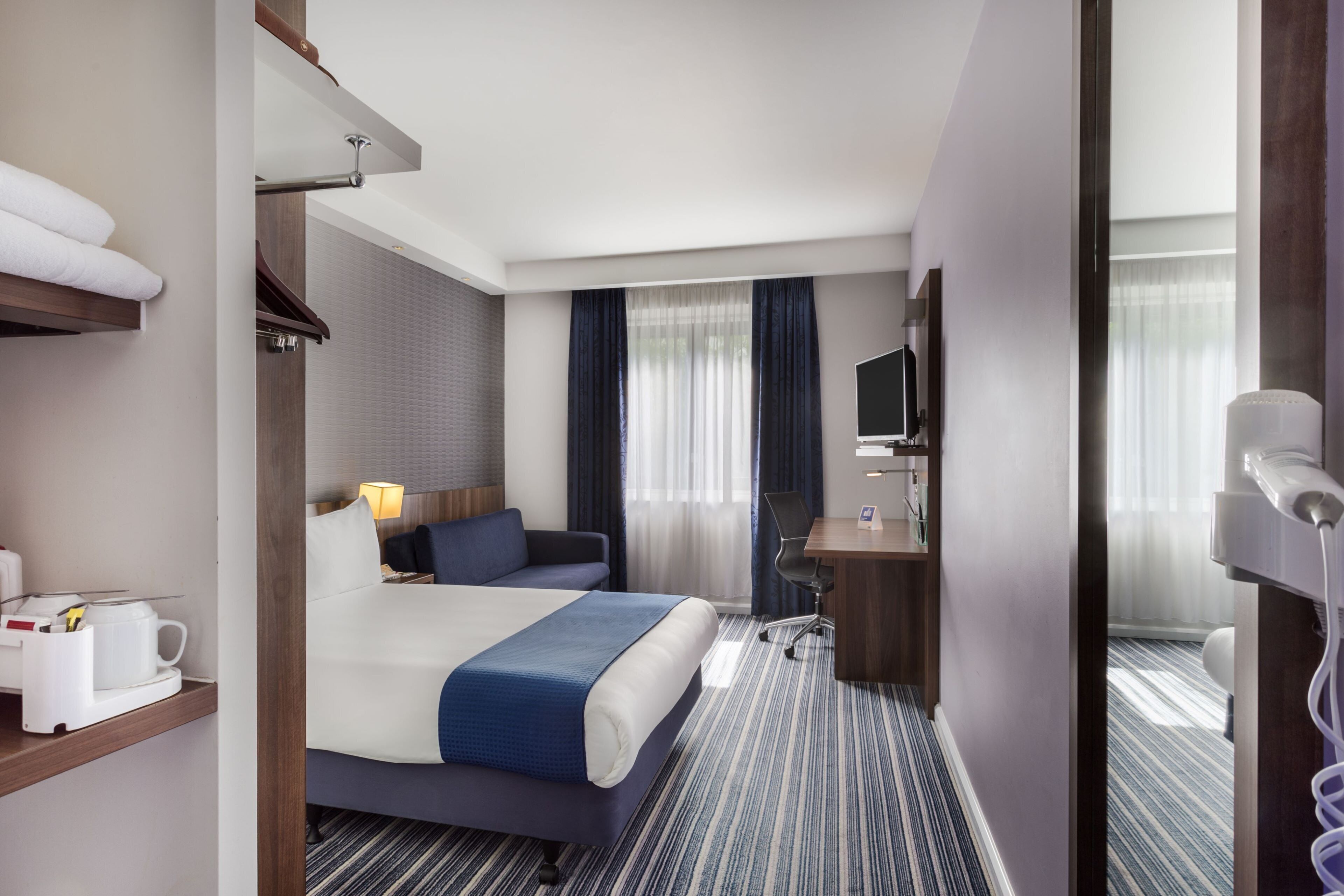 A hotel room with a bed, a desk, a chair, a TV, and a hairdryer.
