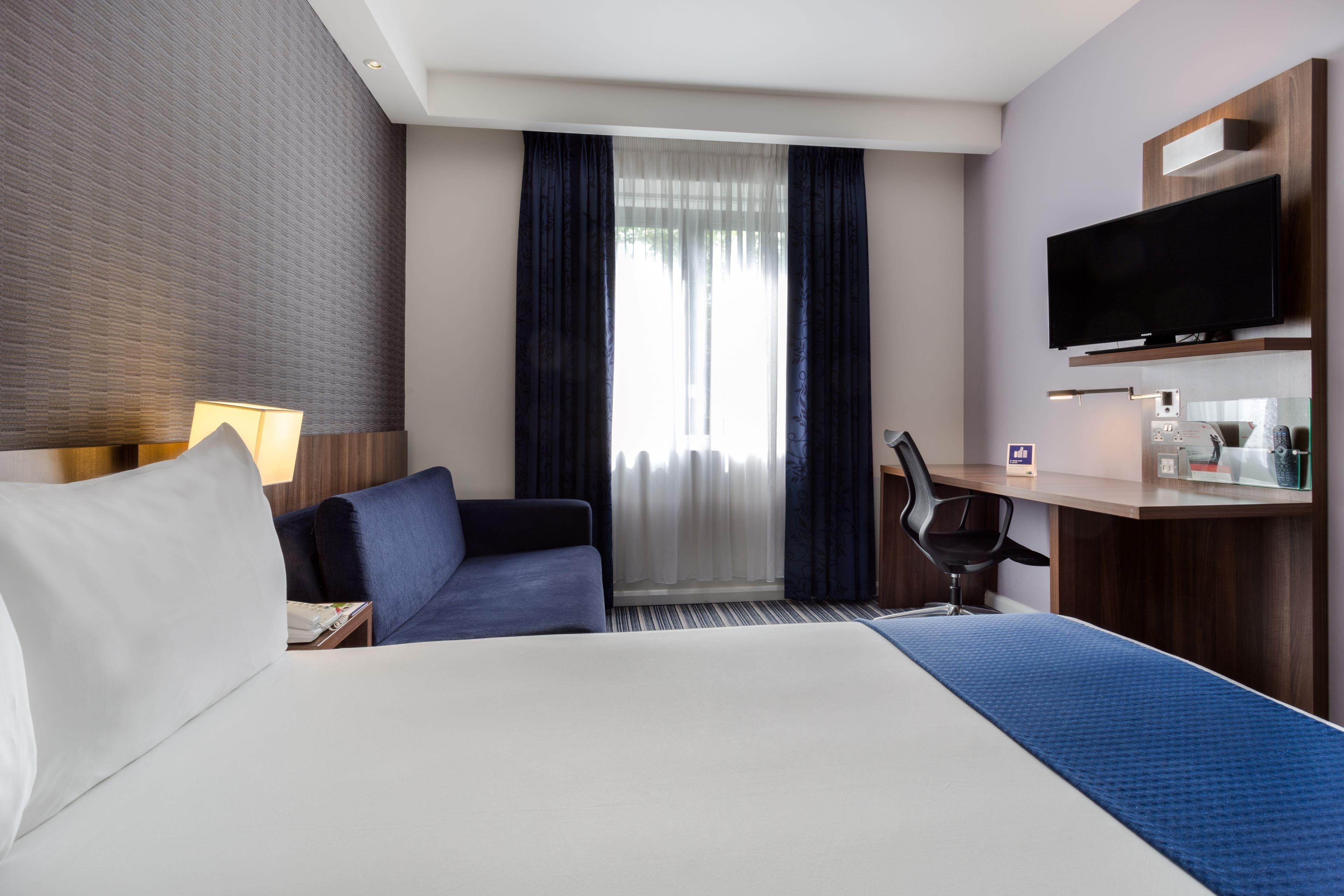 Une chambre d’hôtel avec un lit, un bureau, une chaise, une télévision et une fenêtre avec des rideaux.