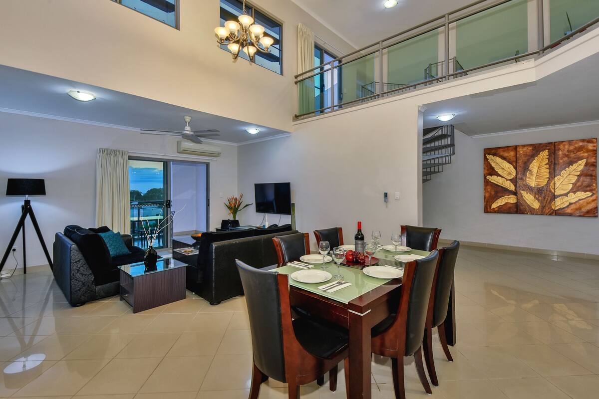A spacious dining area with a high ceiling, a balcony, and a modern chandelier.