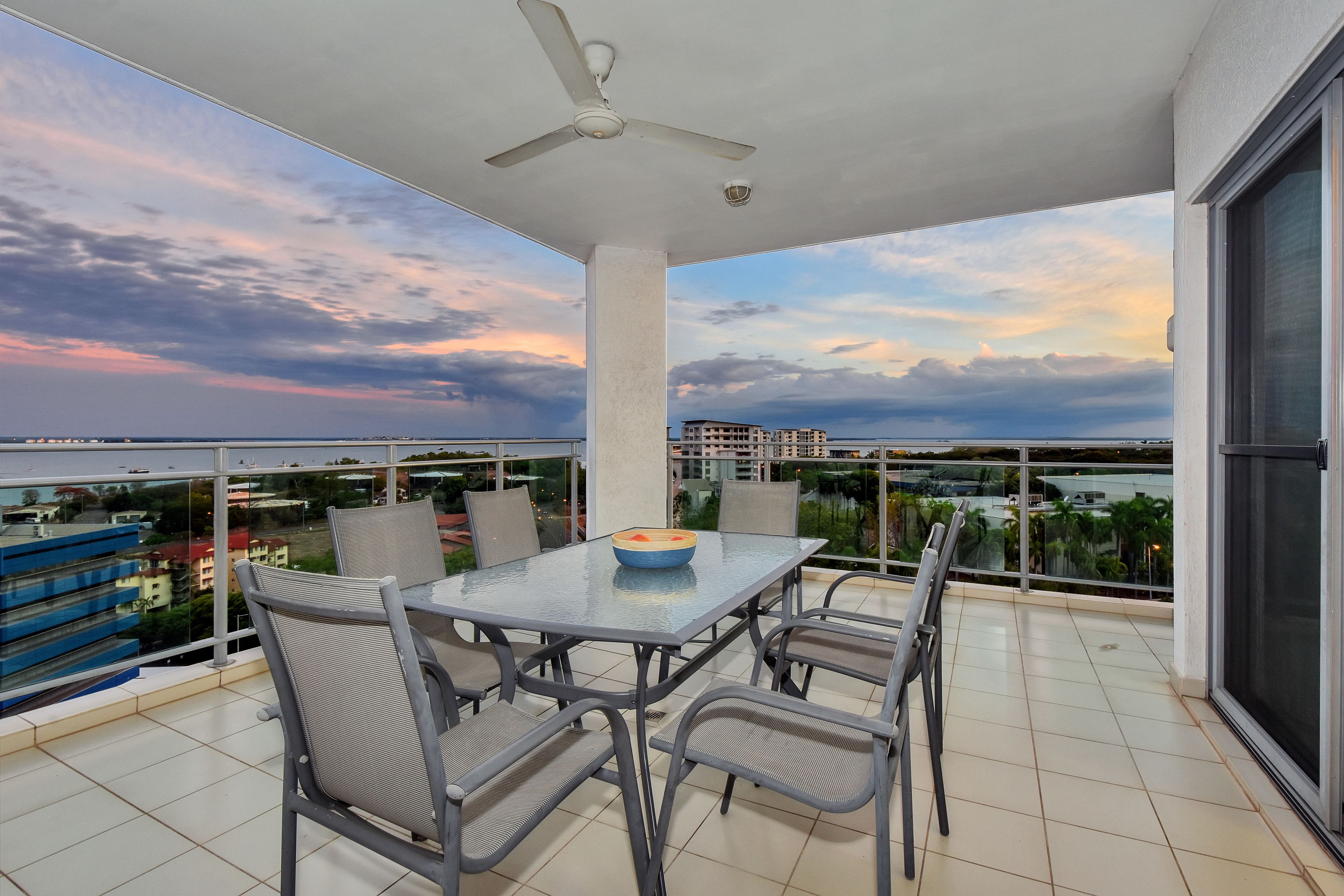 A balcony with a table and chairs, a ceiling fan, and a view of buildings and a body of water.