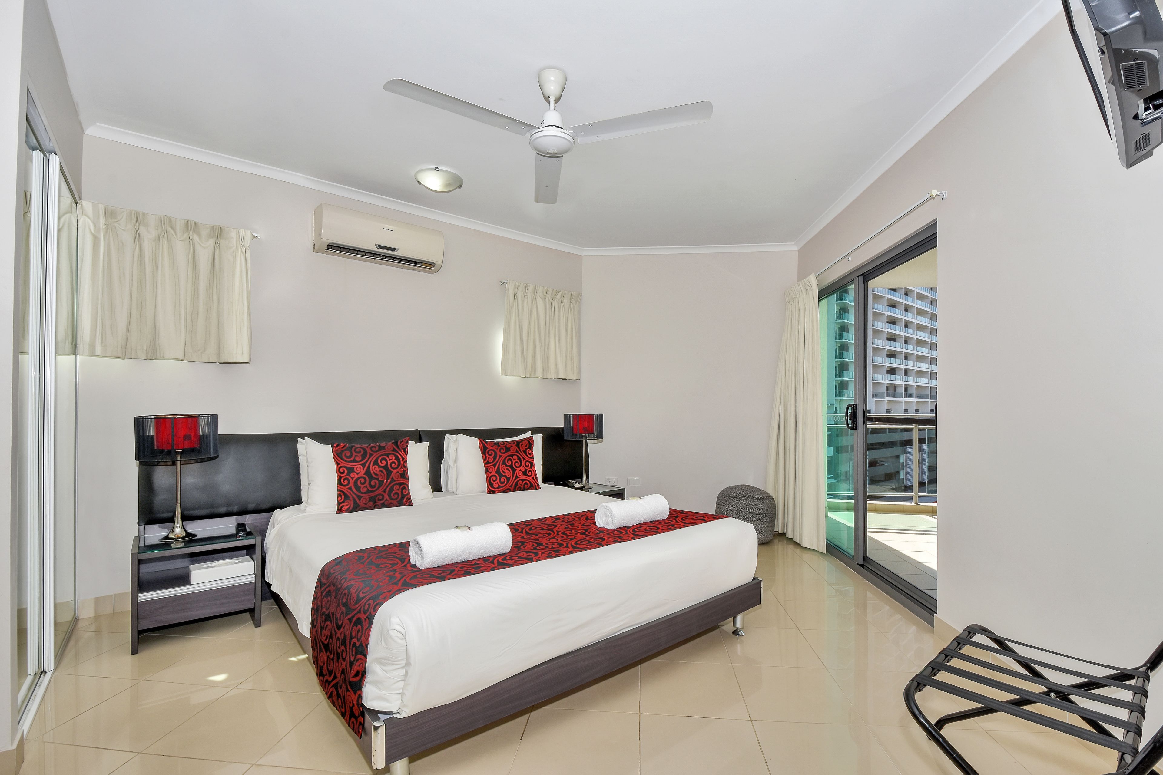 A hotel room with a bed, a nightstand, a ceiling fan, an air conditioning unit, and a view of a building through a glass door.