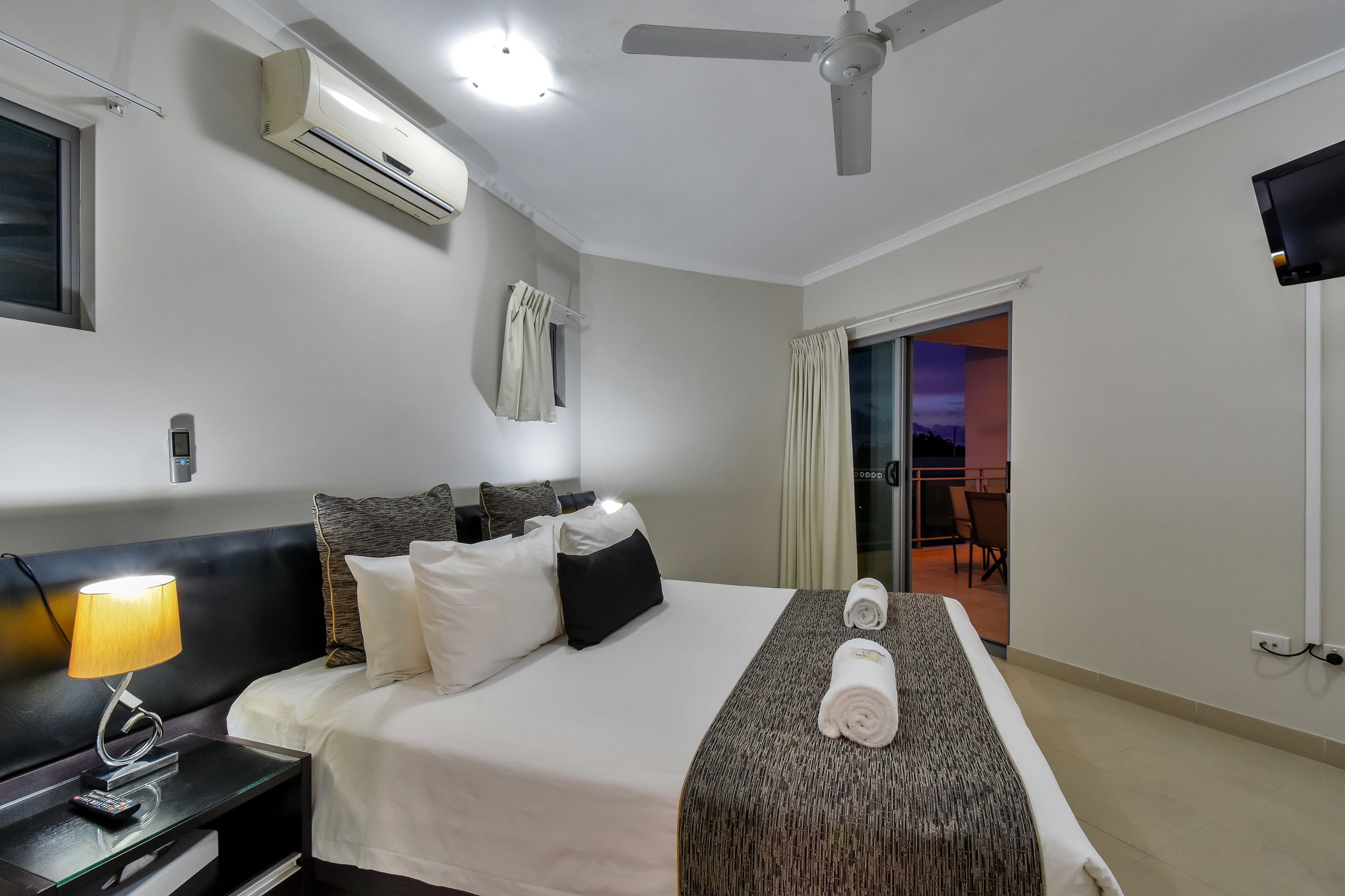 A hotel room with a bed, bedside lamp, air conditioning unit, ceiling fan, and a TV mounted on the wall.