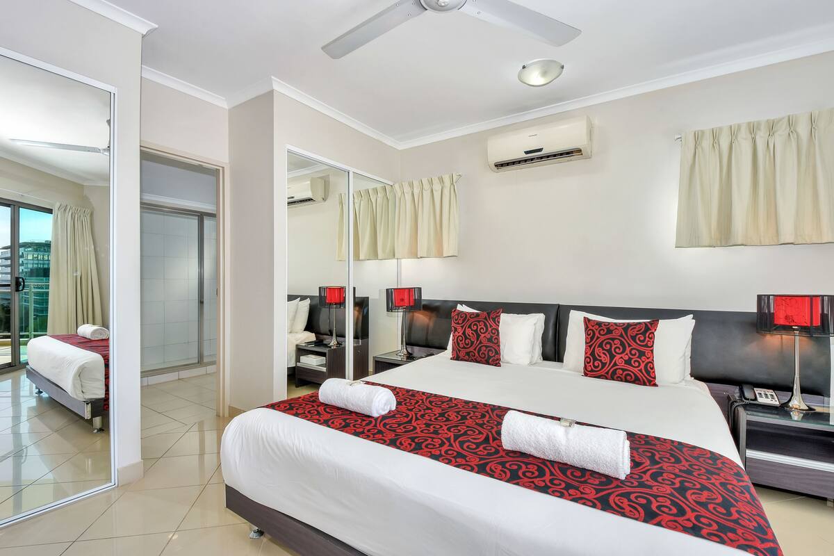 A hotel room with a large bed, a bedside table with a lamp, a mirror, and an air conditioning unit on the wall.