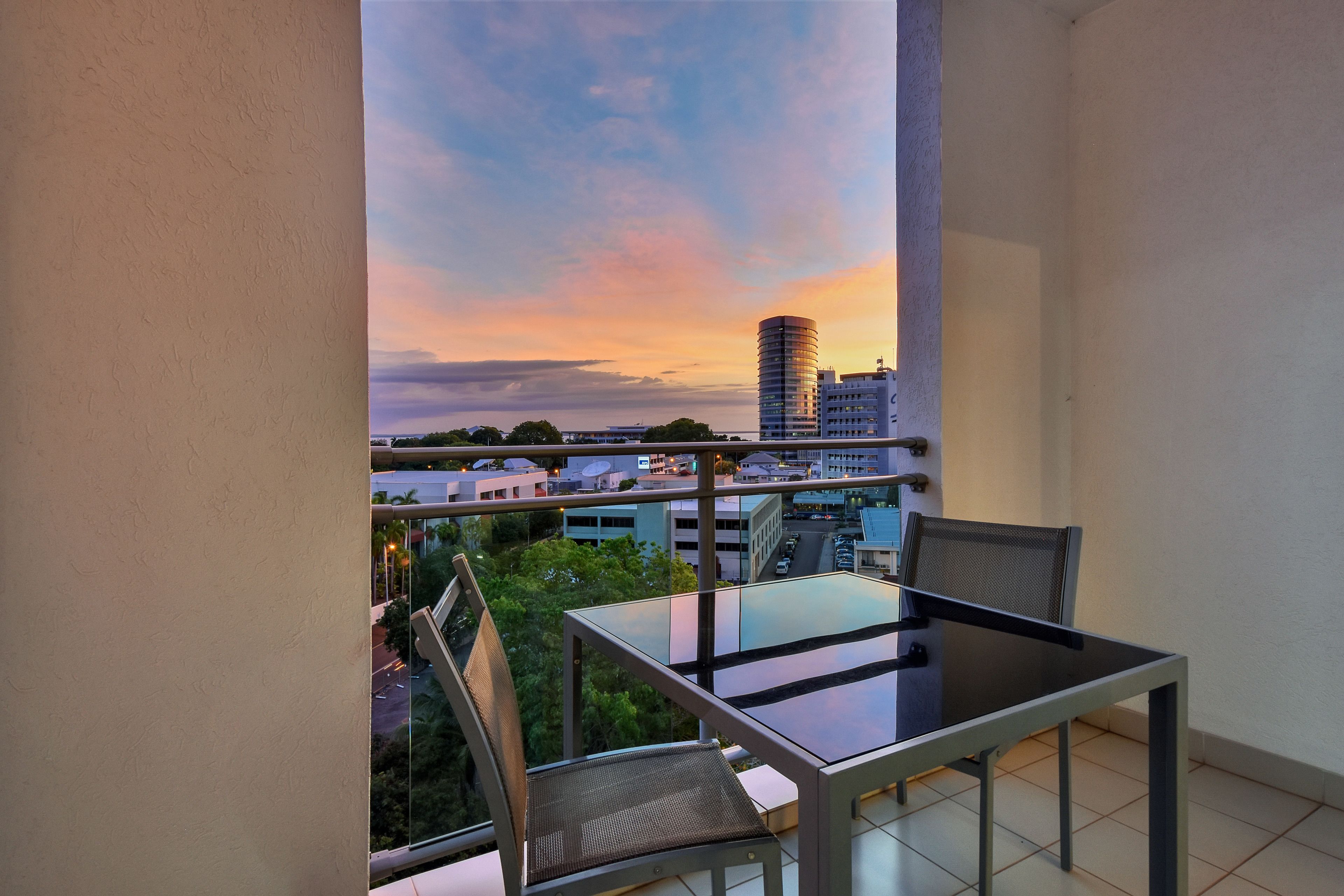 A balcony with a glass table and chairs, overlooking a cityscape at sunset.