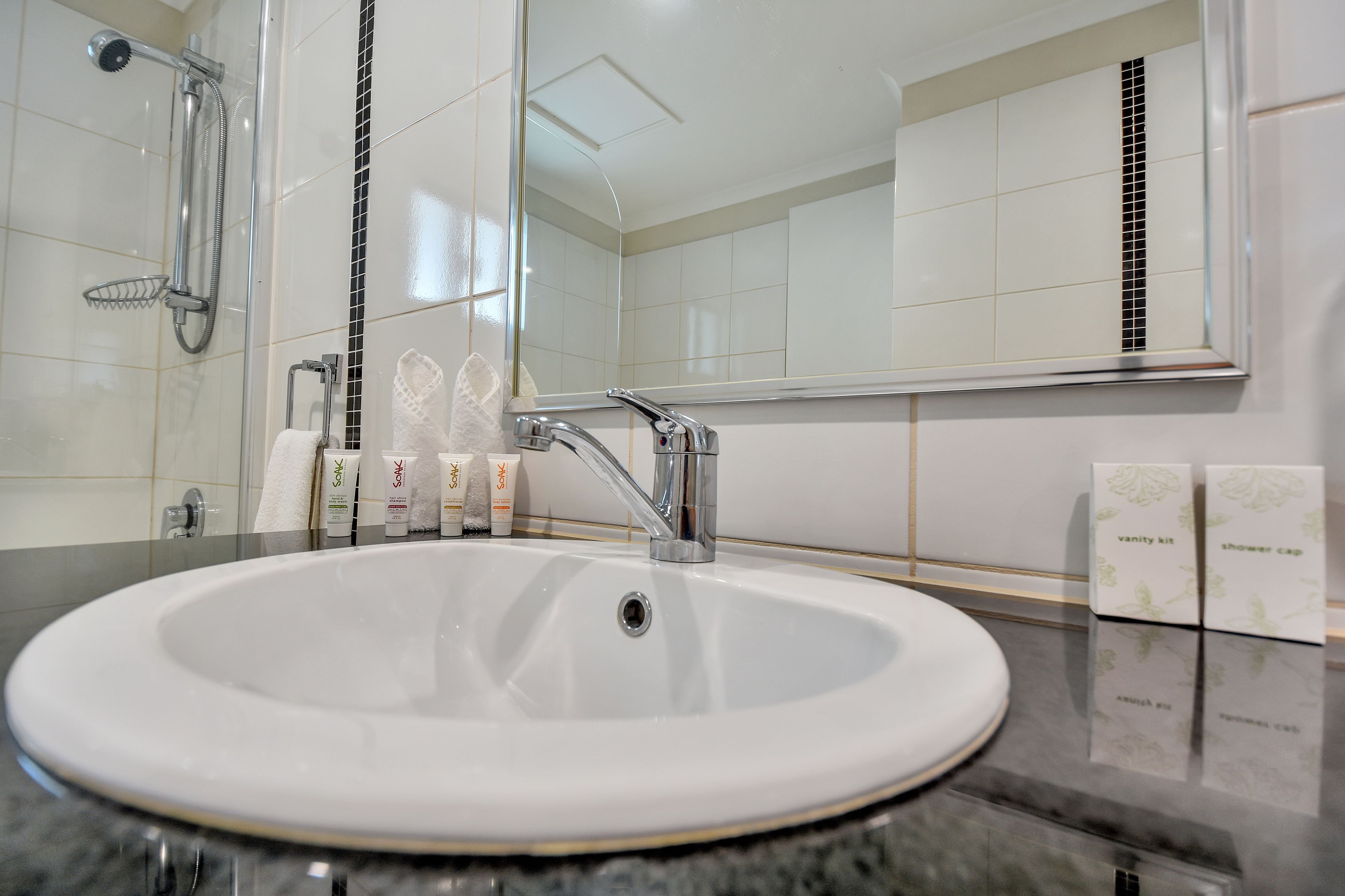 A bathroom with a white sink, chrome faucet, and a showerhead. There are toiletries on the counter and a vanity kit on the wall.