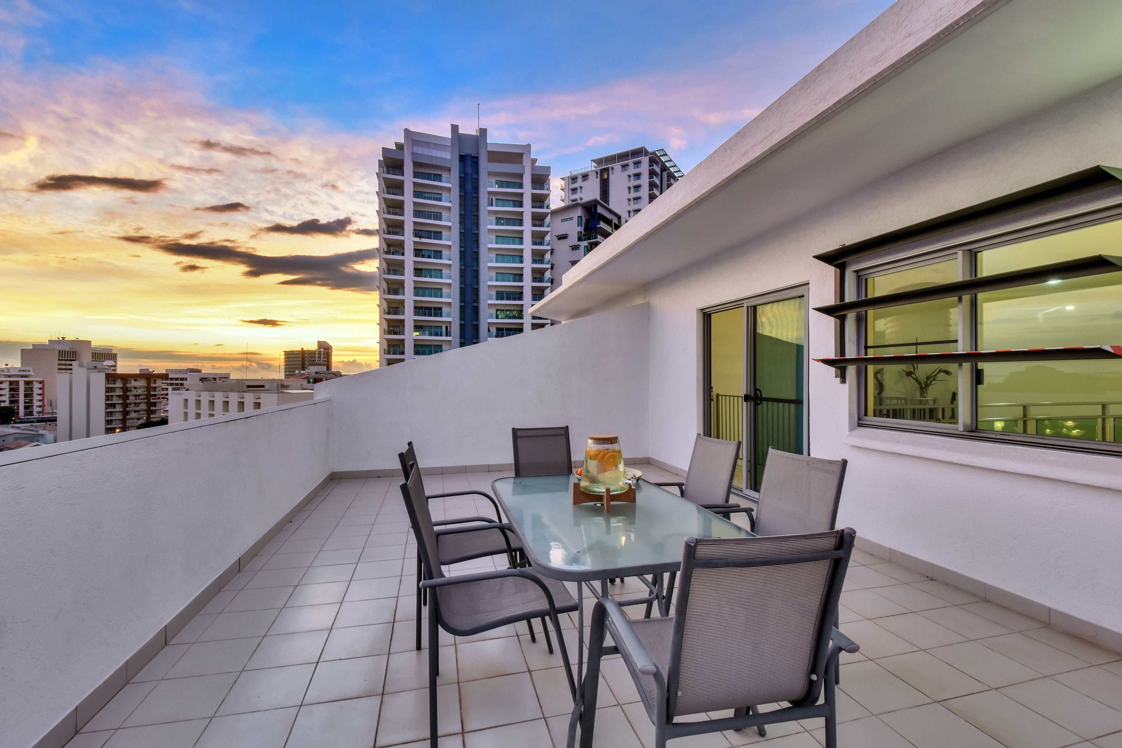 Een balkon met een glazen tafel en stoelen, een decoratieve vaas, en uitzicht op stadsgebouwen tijdens de zonsondergang.