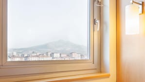 A room with a window overlooking a cityscape and mountains, a wooden table with a cup, and a wall-mounted light fixture. - ibis Kayseri (Kocasinan)