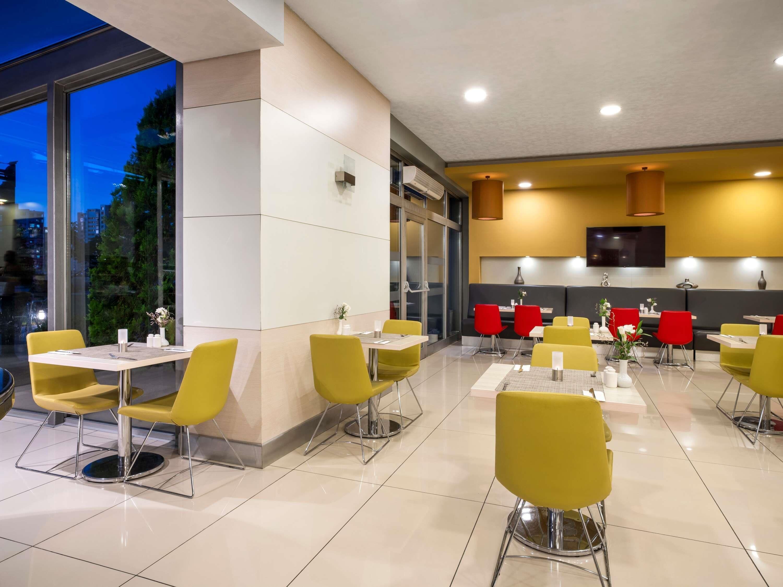 Un restaurant modern amb cadires grogues i vermelles, un televisor de pantalla plana i vista a la ciutat a l'hora del vespre.