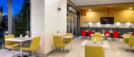 A modern restaurant with yellow and red chairs, a flat-screen TV, and a view of the city at dusk.