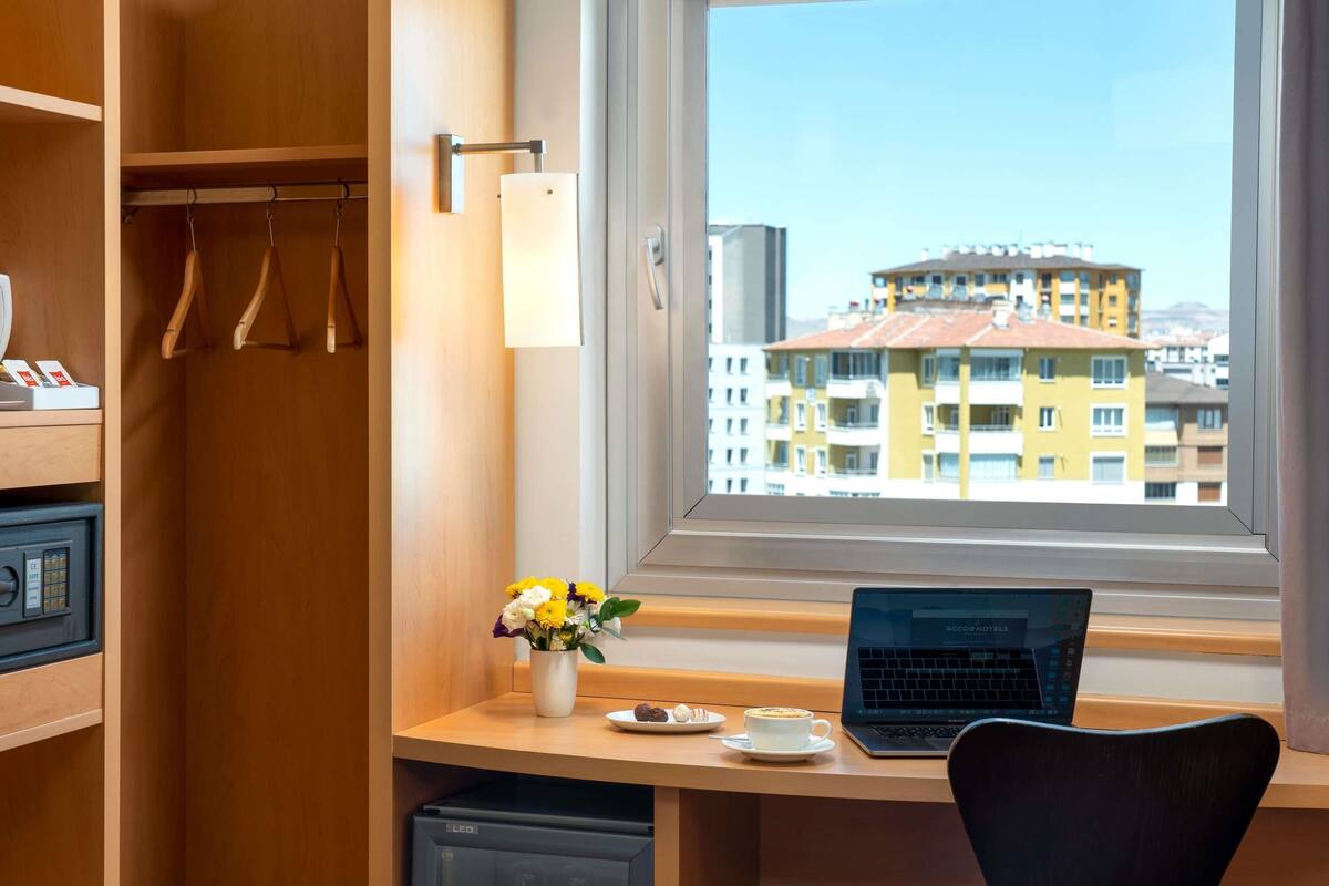 A hotel room with a wooden desk, a laptop, a coffee cup, a small fridge, a vase with flowers, and a view of buildings through a window.