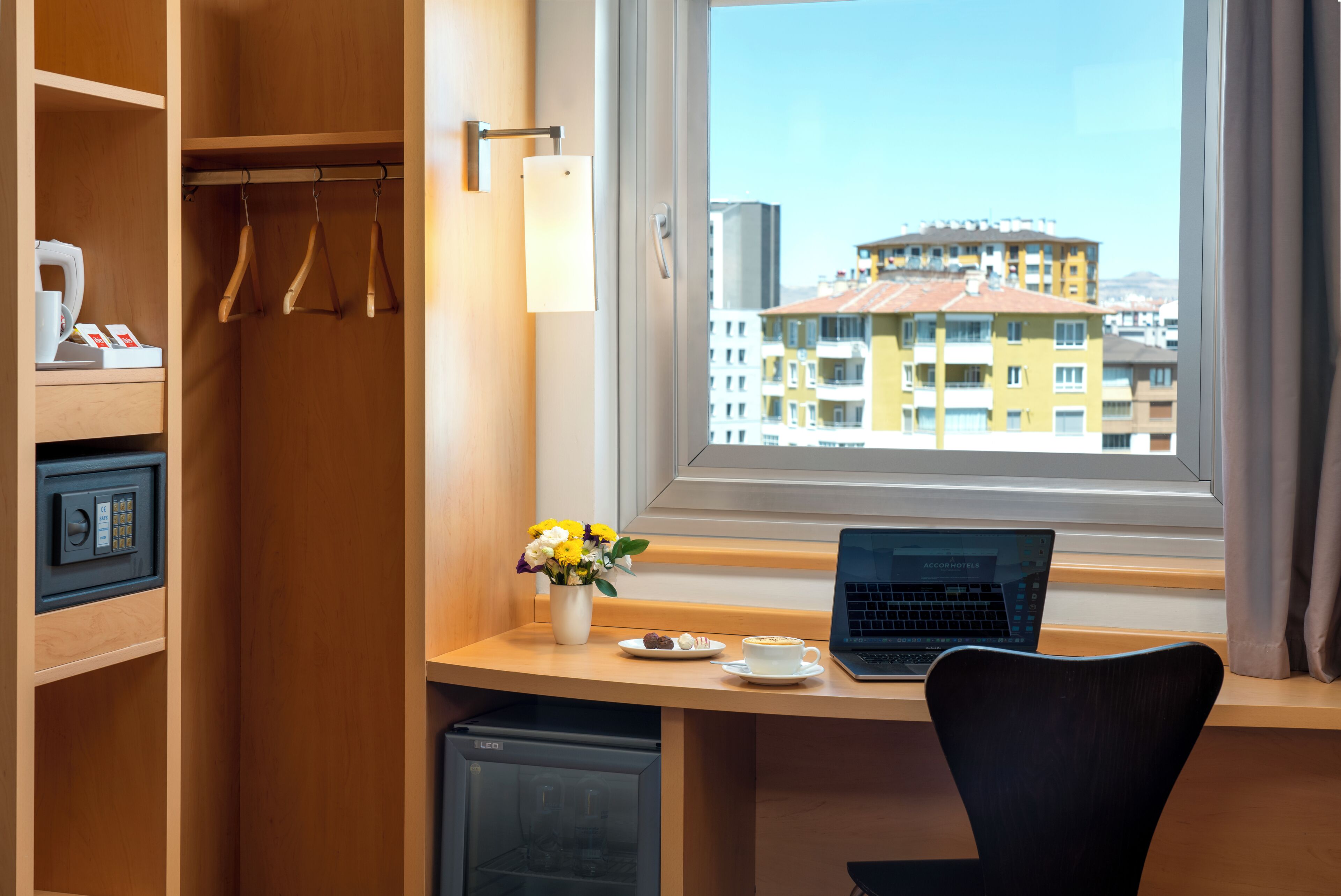 A hotel room with a desk, laptop, coffee cup, and a view of buildings.