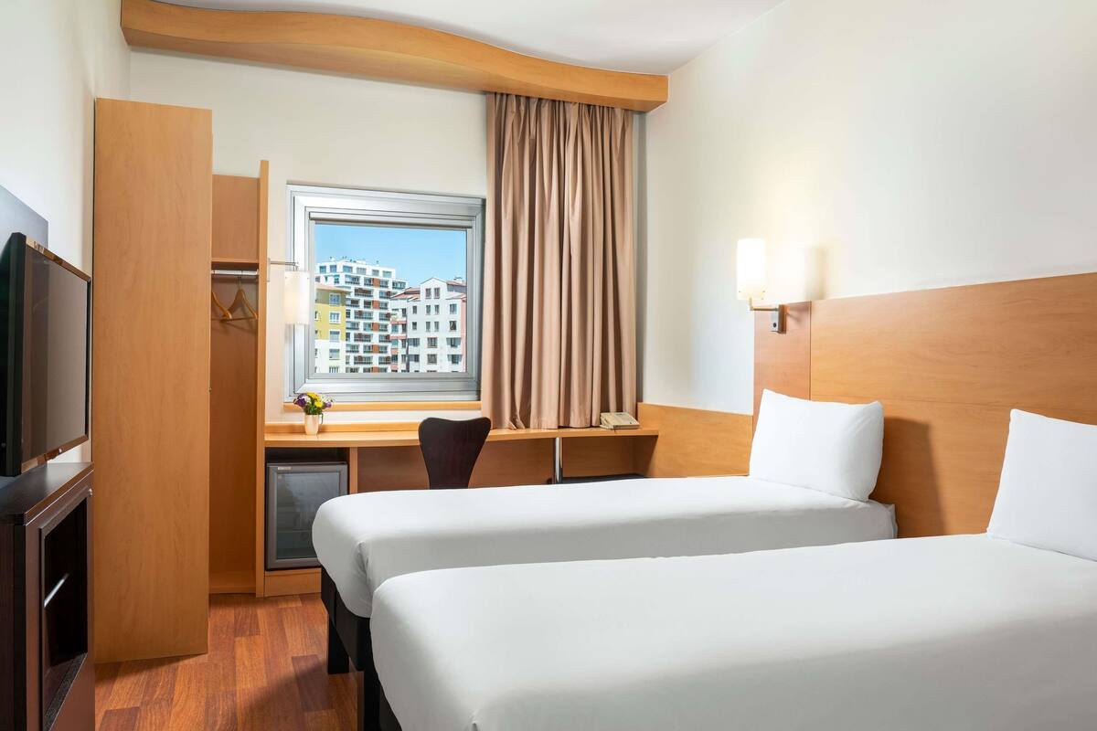 A hotel room with two beds, a desk with a chair, a TV, and a window with a view of buildings.