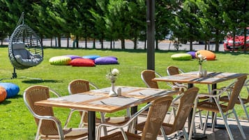 An outdoor seating area with tables and chairs, surrounded by greenery and colorful cushions.