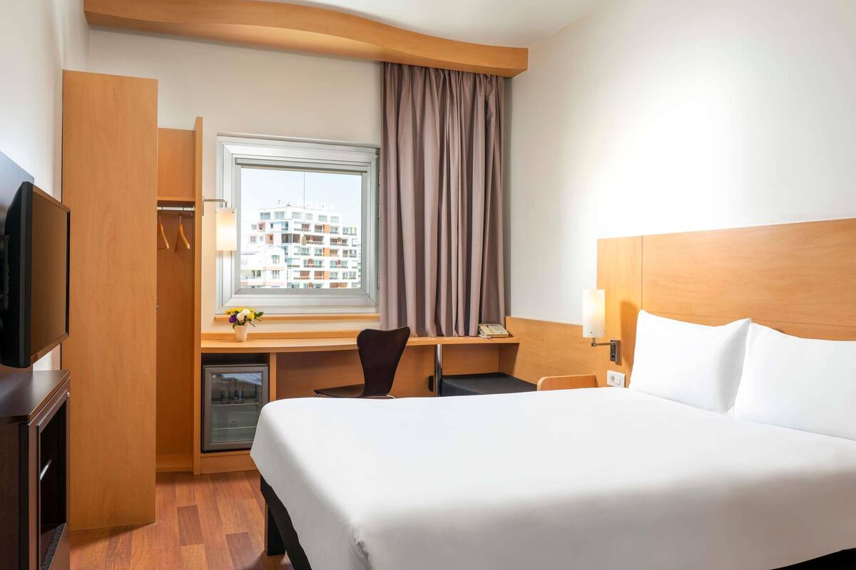 A hotel room with a bed, a desk with a chair, a TV, a window with curtains, and a view of buildings outside.