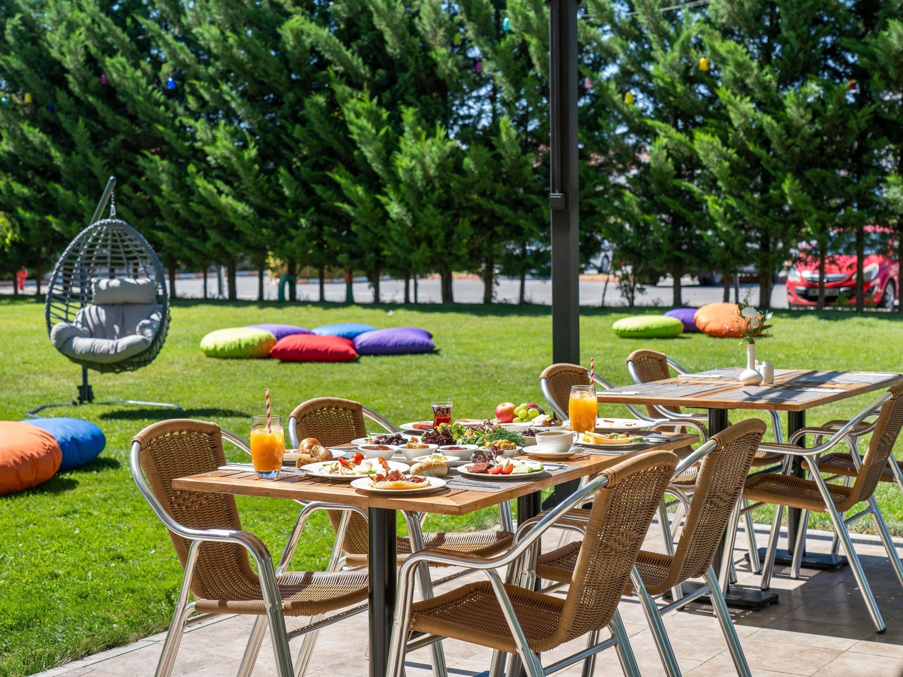 Un espace de restauration en plein air, avec des chaises et des tables en osier, entouré de pelouse verdoyante et d’arbres.