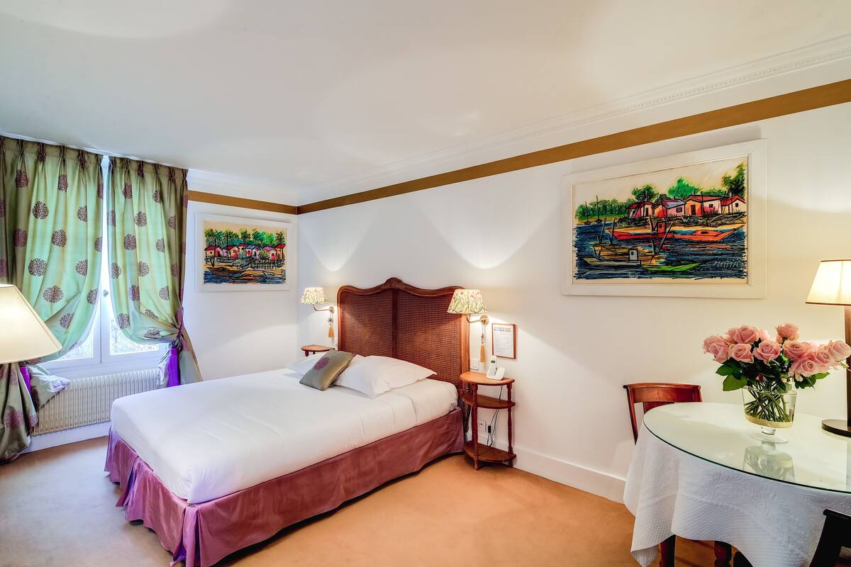 A hotel room with a bed, a bedside table with lamps, a chair, a glass table with a vase of flowers, and two paintings on the wall.