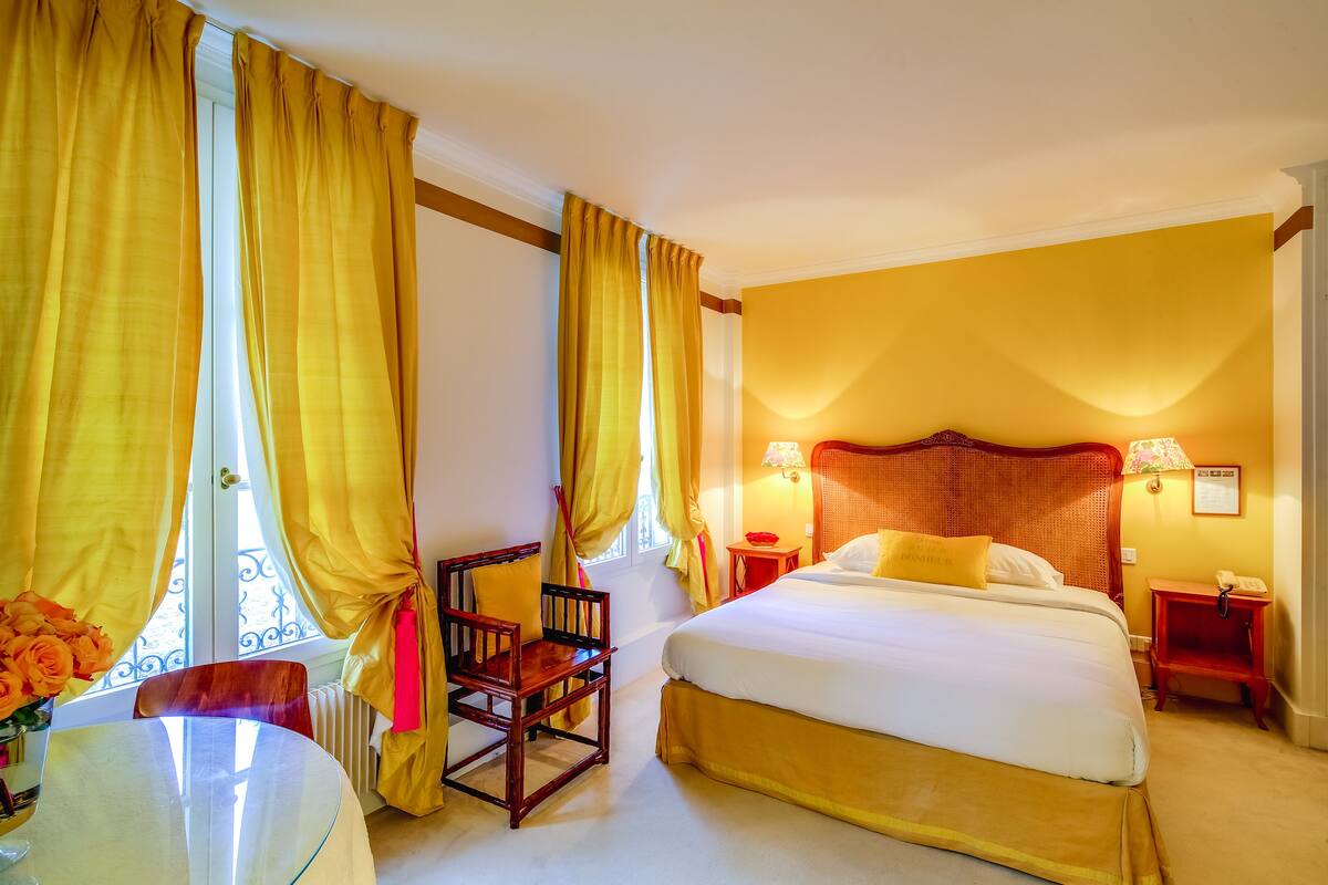 A hotel room with a large bed, a wooden chair, a small table with a vase of flowers, and a window with yellow curtains.