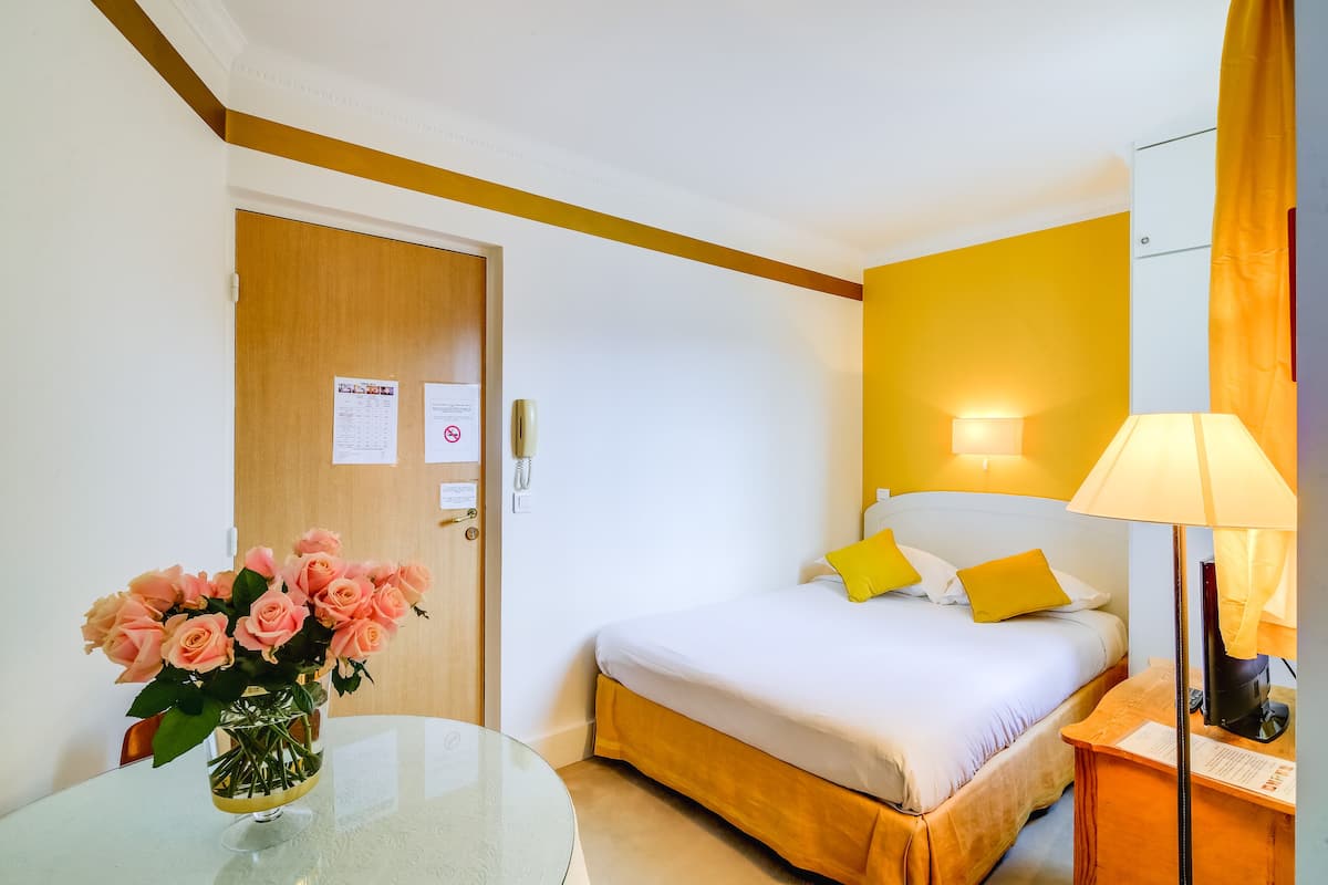 A hotel room with a bed, a bedside table, a lamp, a vase with flowers, and a door with a bulletin board.