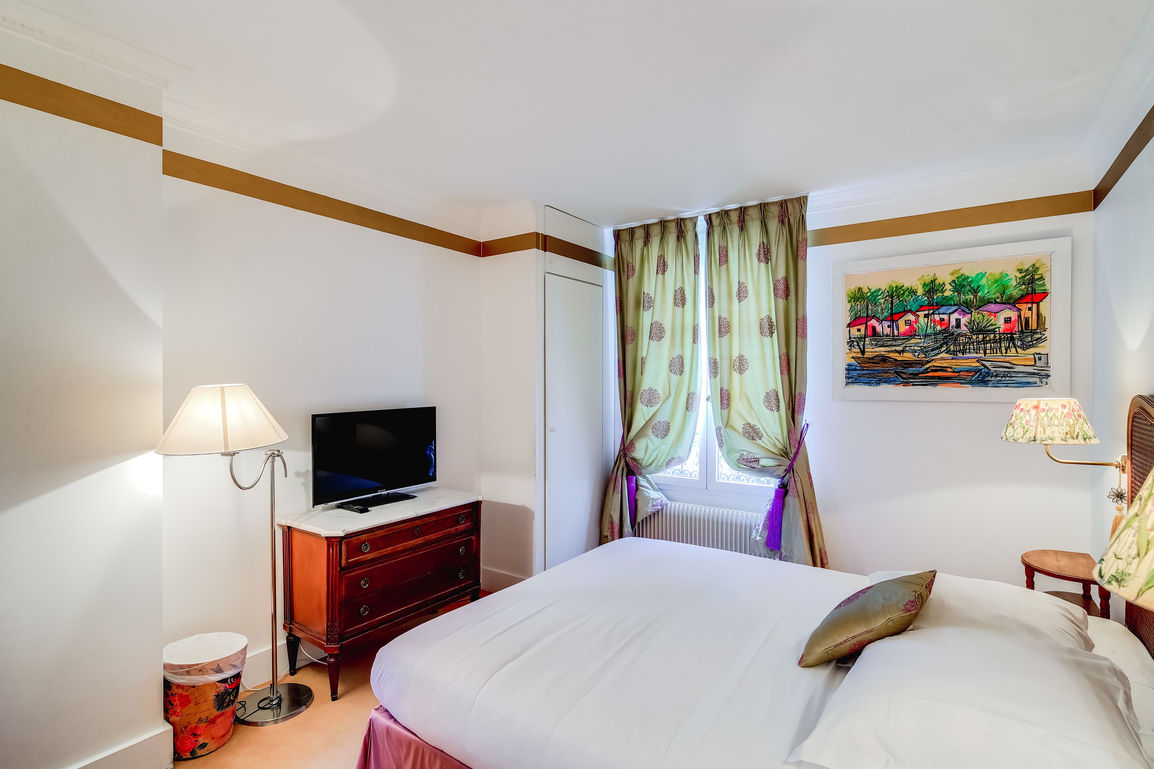 A hotel room with a bed, a television on a dresser, a lamp, a painting on the wall, and a window with curtains.