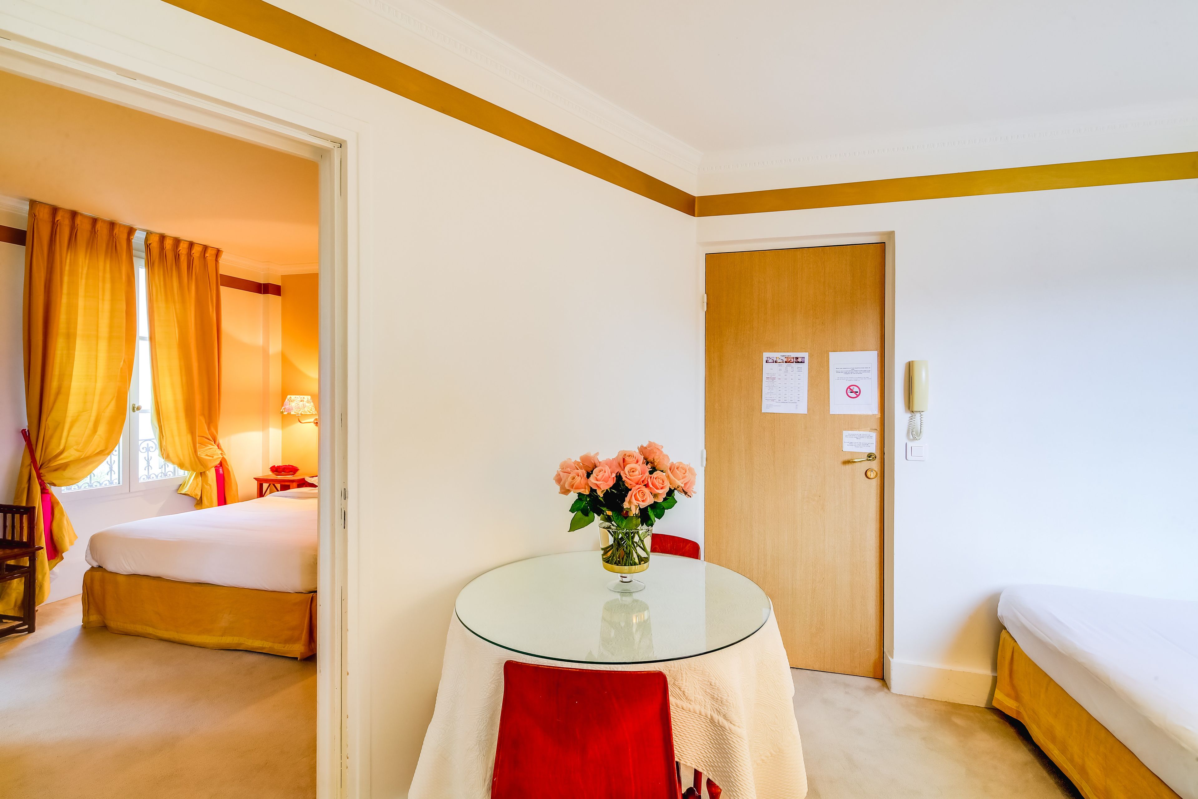 Ett hotellrum med en säng, ett litet bord med en vas med blommor, en dörr med en skylt och ett fönster med gardiner.