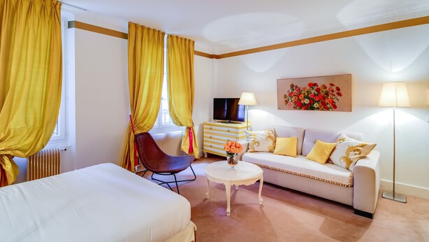 Habitación de hotel con sofá, una silla, una mesita con flores, televisor y radiador.