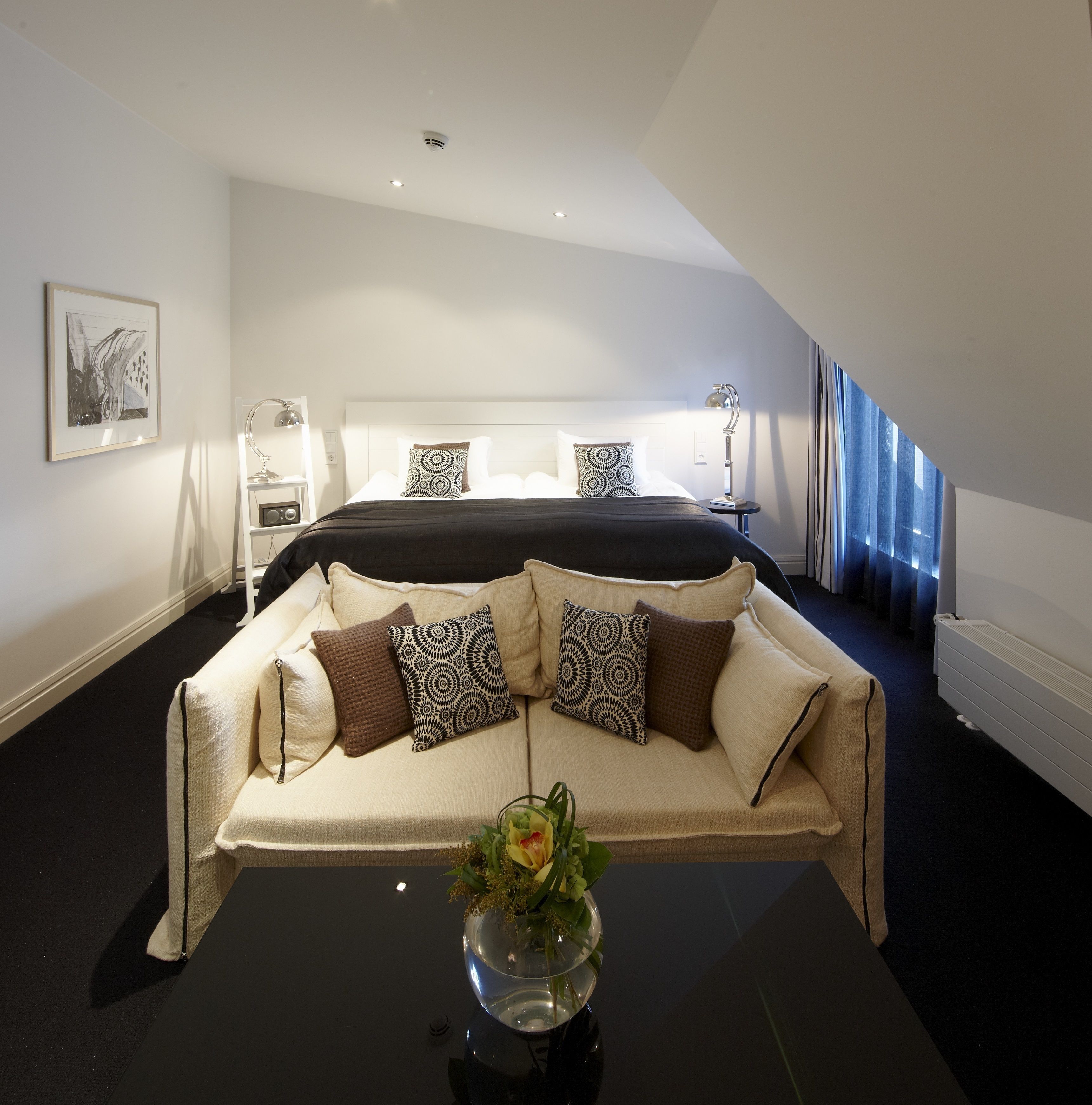 Una habitación de hotel moderna con una cama grande, un sofá, una mesita de noche con lámpara, un cuadro en la pared y una ventana con cortinas.