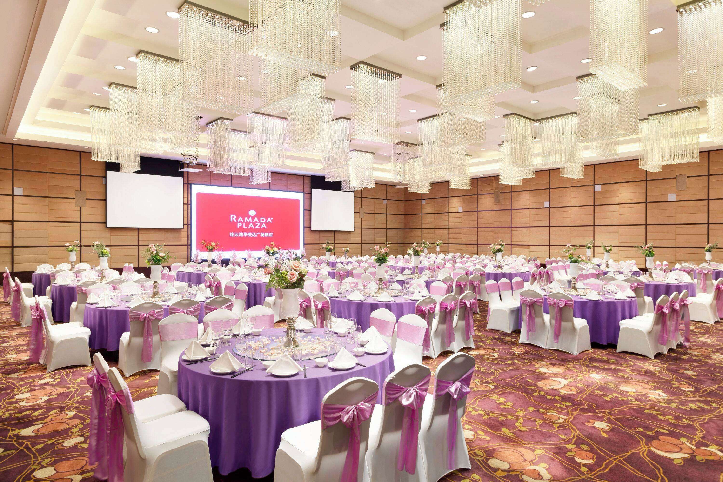 A large banquet hall with round tables covered in purple tablecloths, white chairs with pink sashes, and a large screen displaying "RAMADA PLAZA".
