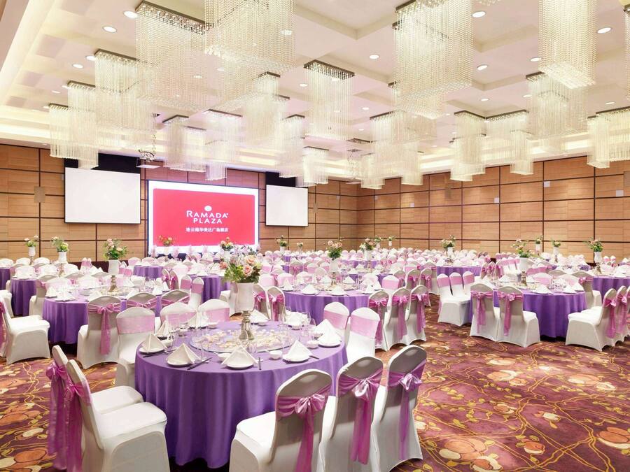 A large banquet hall with round tables covered in purple tablecloths, white chairs with pink sashes, and a large screen displaying "RAMADA PLAZA".