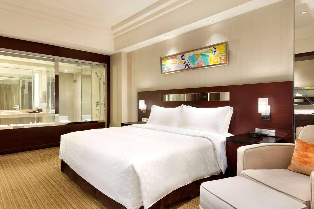 Hotelkamer met een groot bed, een zithoek, een badkamer met douchecabine van glas en een kleurrijk abstract schilderij aan de muur.