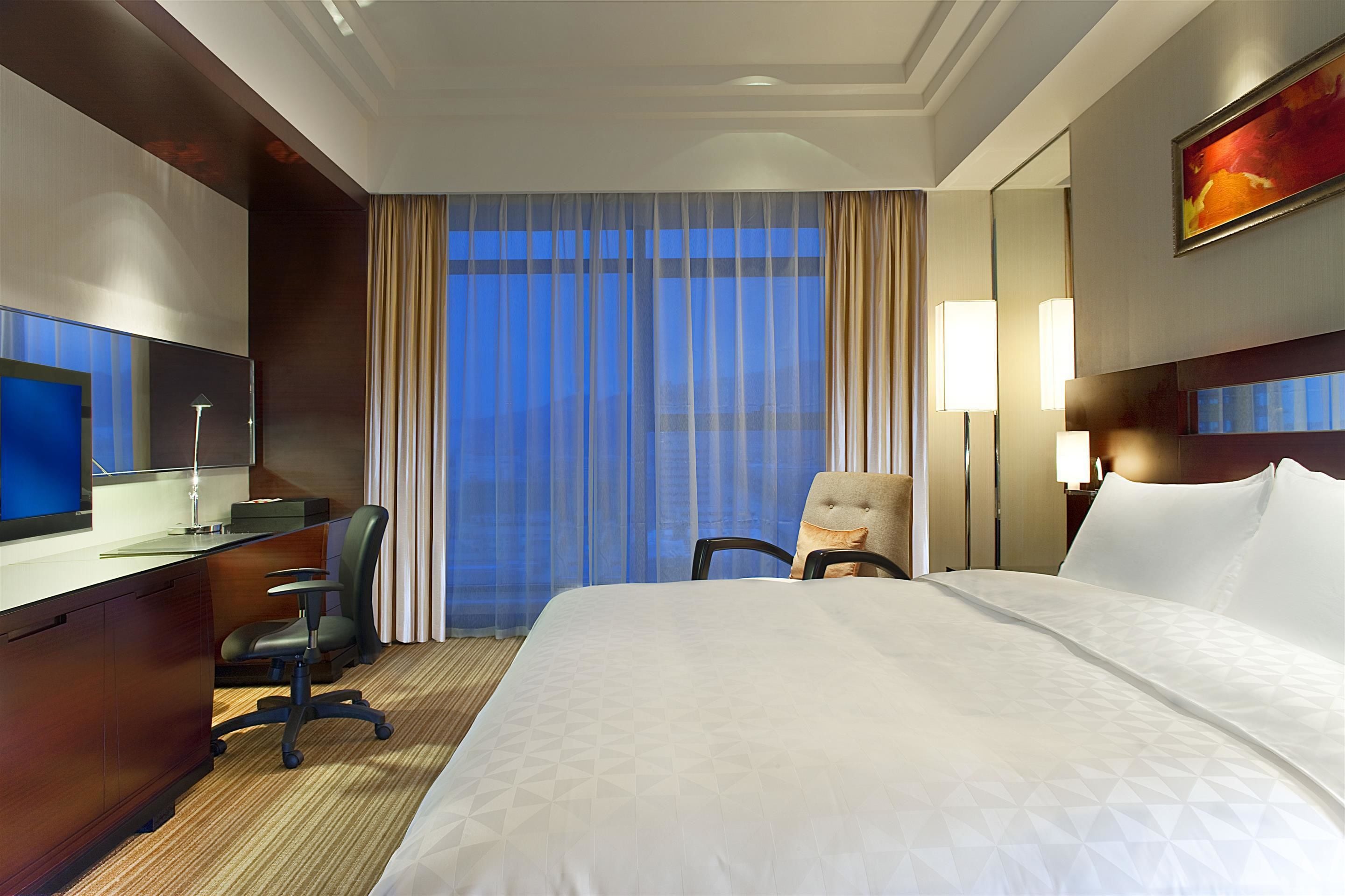 Ein Hotelzimmer mit einem großen Bett, einem Schreibtisch mit Computer, einem Stuhl und Blick auf die Stadt durch transparente Vorhänge.