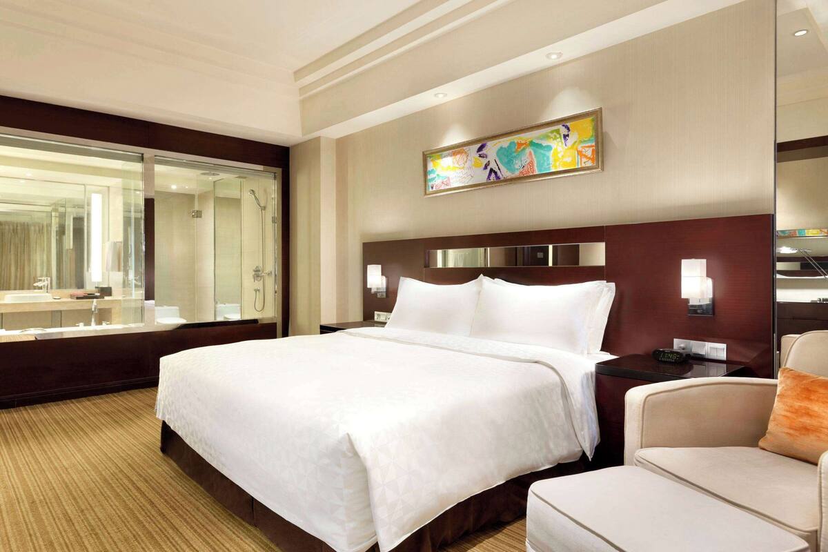 Een hotelkamer met een groot bed, een zithoek met een fauteuil, een grote spiegel en een schilderij aan de muur.