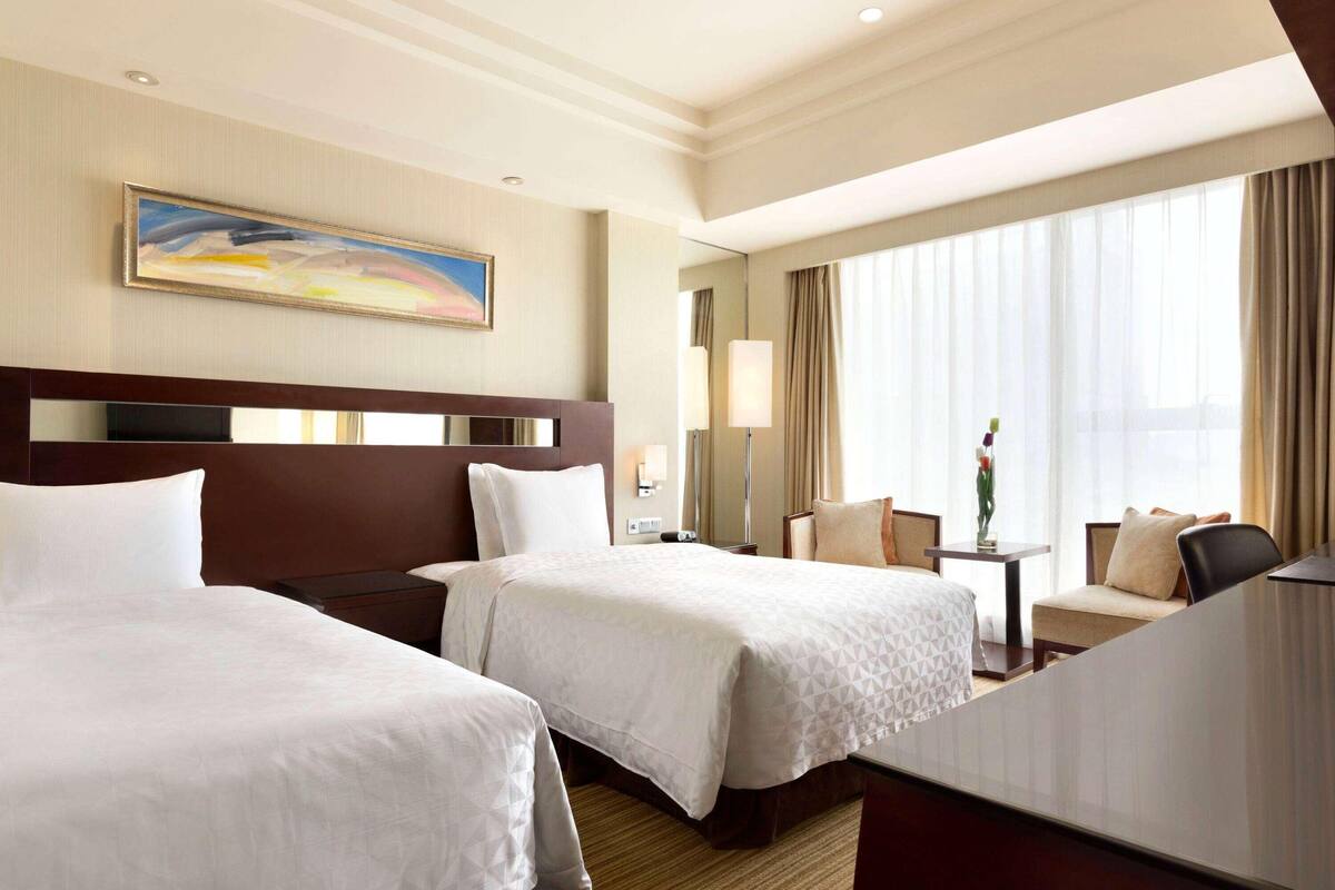 Een hotelkamer met twee bedden, een groot raam met gordijnen, een schilderij aan de muur en een klein tafeltje met een vaas.