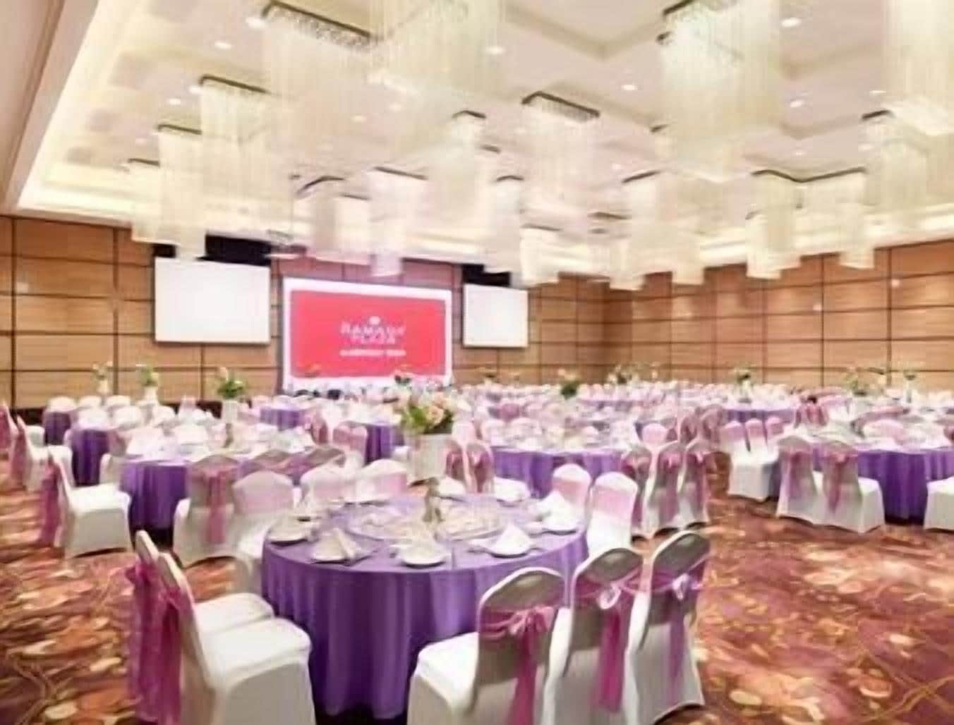 A banquet hall with round tables covered in purple tablecloths, white chairs with pink sashes, and a large screen displaying text.