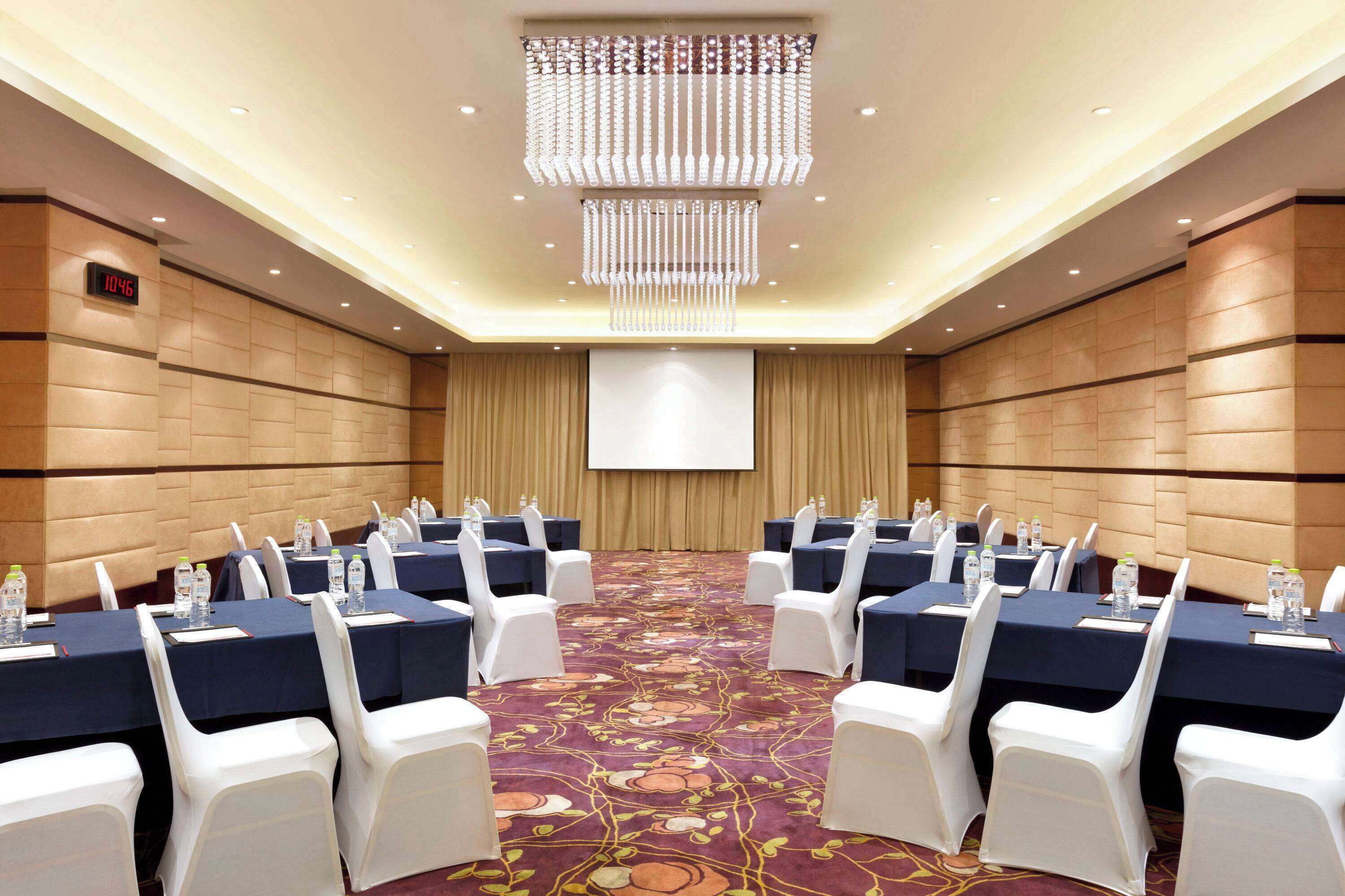 A conference room with rows of tables and chairs, a projector screen, and a chandelier.
