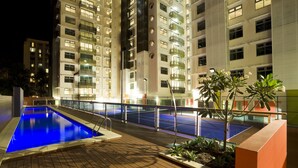 Un espace piscine doté d’une terrasse en bois, entouré d’une rambarde, et situé à côté d’un immeuble résidentiel à plusieurs étages, doté de balcons.