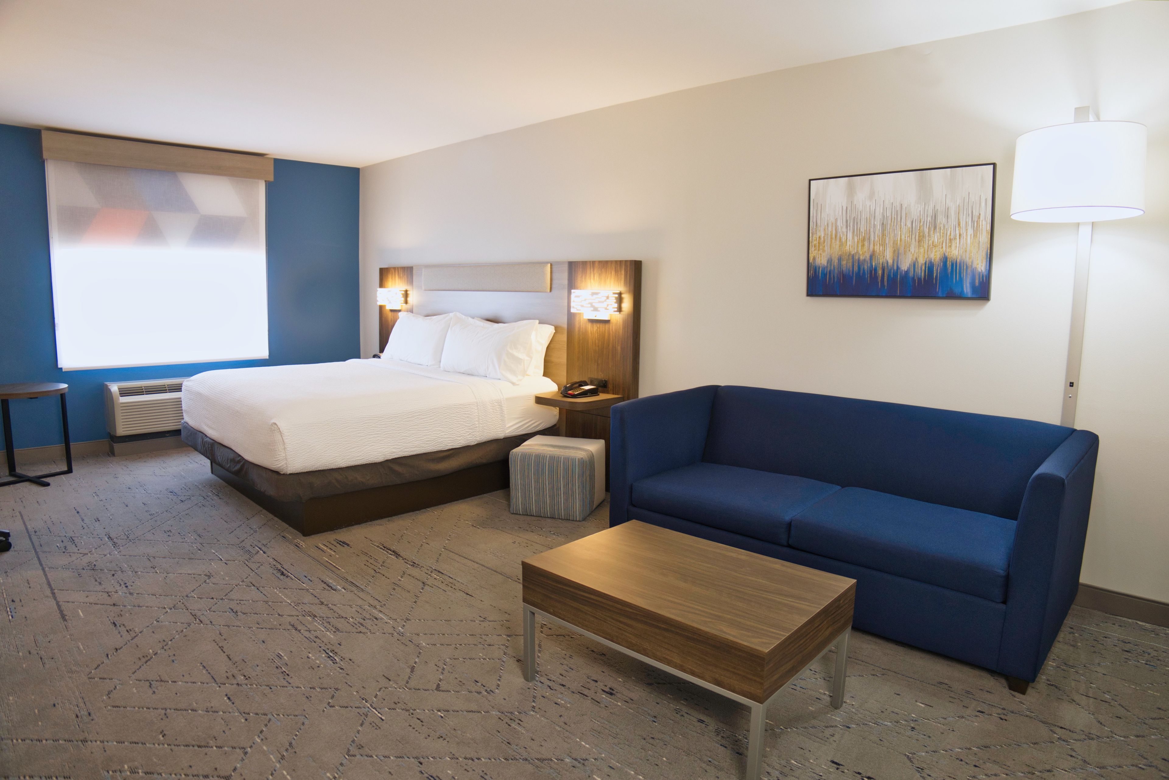 Une chambre d’hôtel avec un lit, un canapé bleu, une table basse en bois, une table de chevet, une lampe et un tableau au mur.