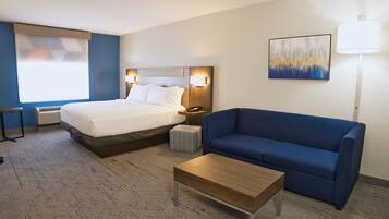 Une chambre d’hôtel avec un lit, un canapé bleu, une table basse en bois, une table de chevet, une lampe et un tableau au mur.