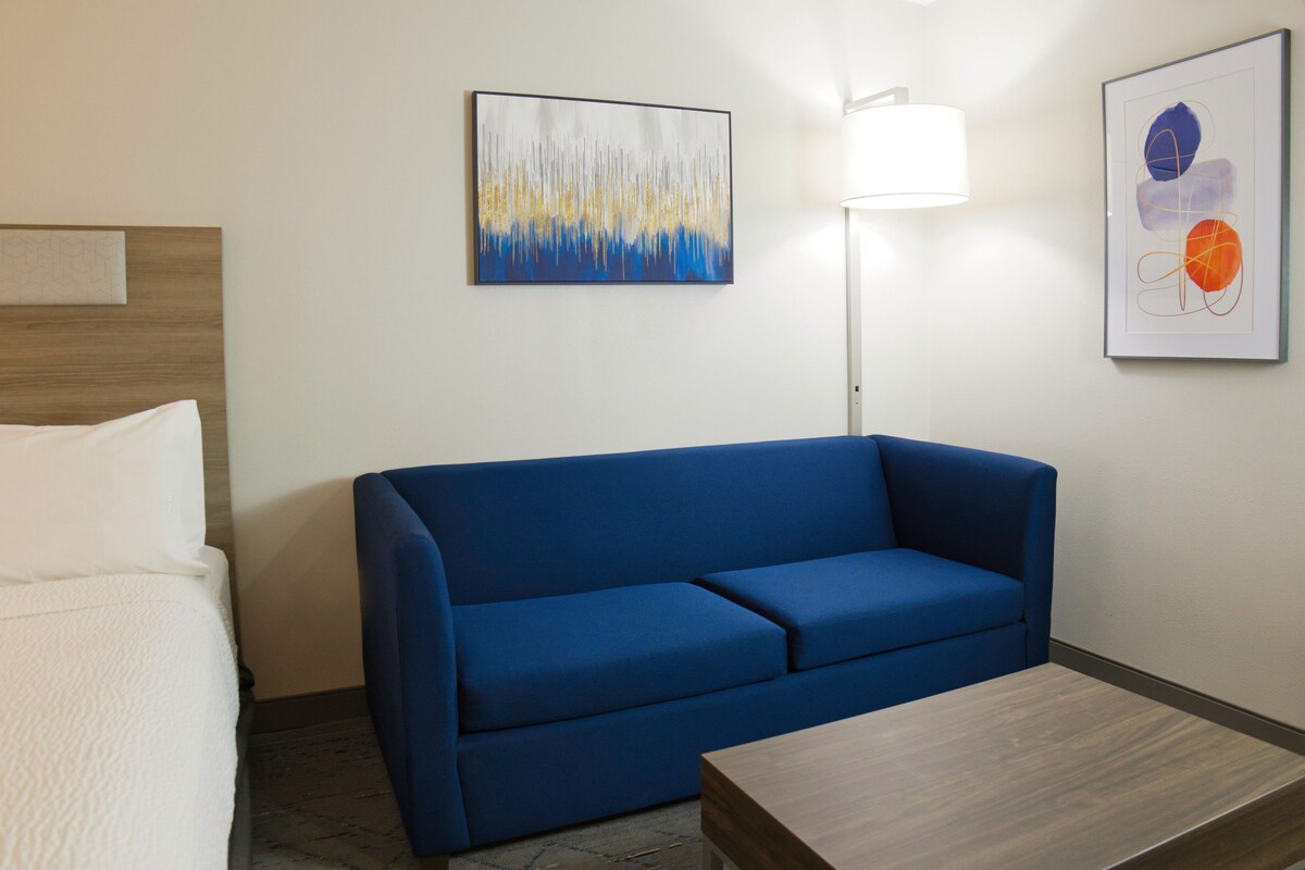 A hotel room with a blue sofa, a wooden table, a lamp, and two paintings on the wall.