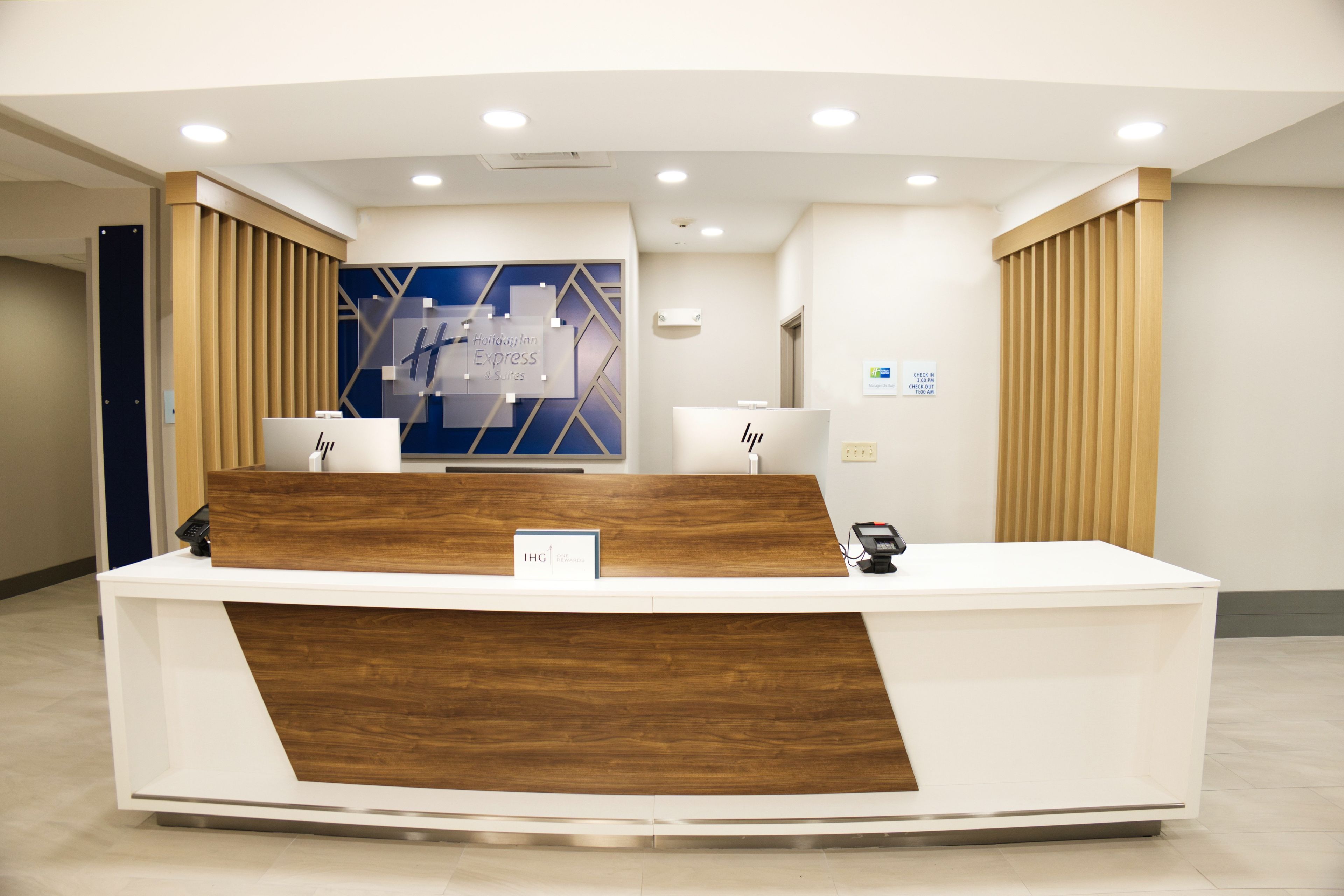 A hotel reception desk with a modern design, featuring a wooden accent panel, a digital check-in kiosk, and a blue wall with geometric patterns in the background.