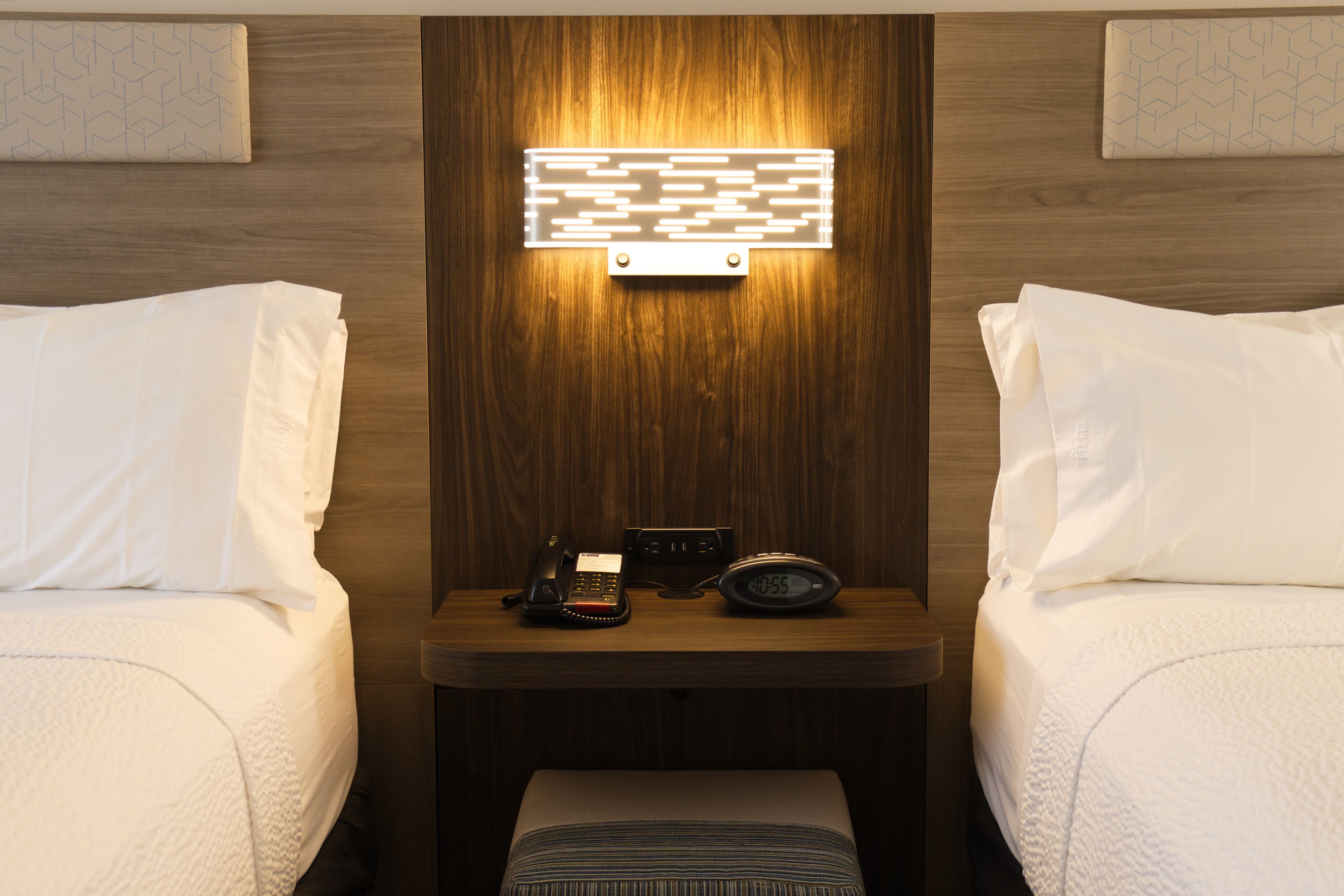 A hotel room with two beds, a bedside table with a phone and an alarm clock, and a wall-mounted light fixture.