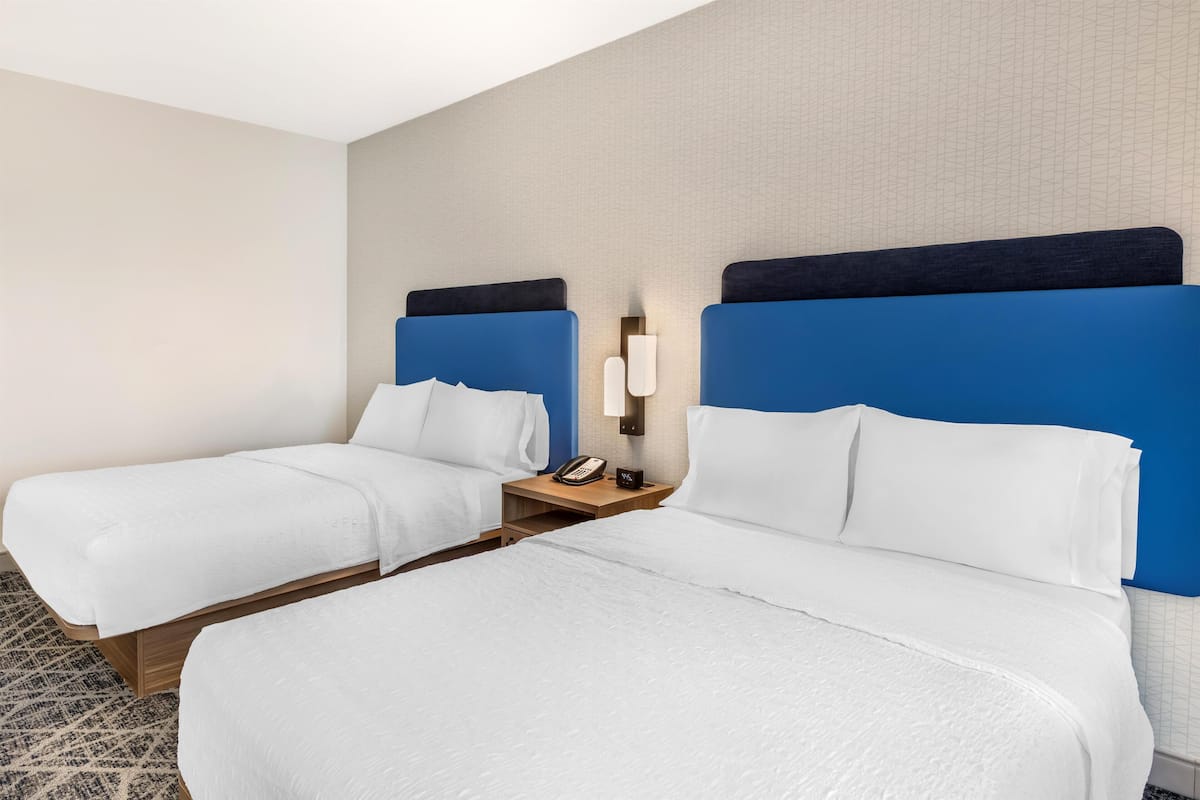 A hotel room with two beds, a nightstand with a phone, and a wall-mounted light fixture.