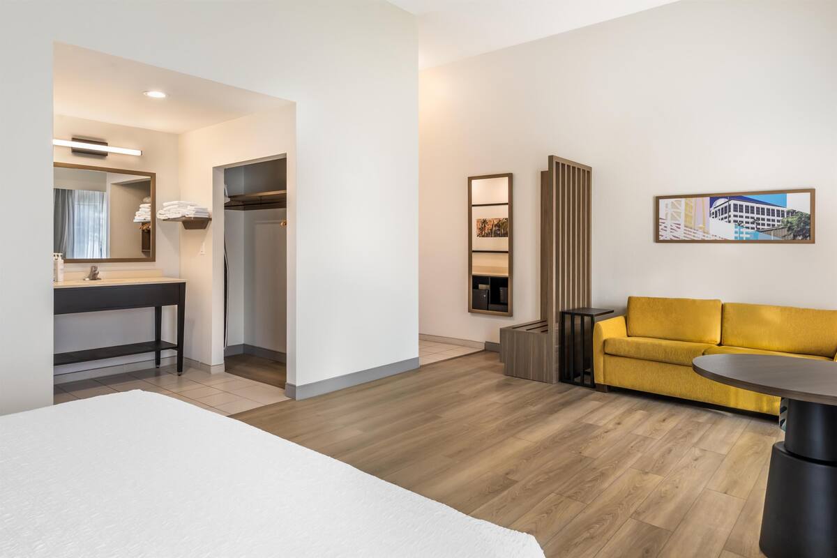 A hotel room with a bed, a bathroom with a mirror and sink, a yellow sofa, and a small table.