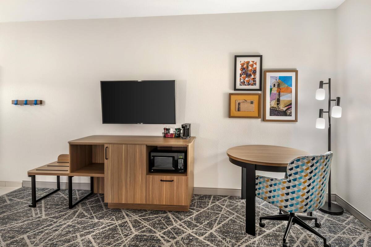A hotel room with a wooden desk, a microwave, a TV, a patterned chair, and a floor lamp.