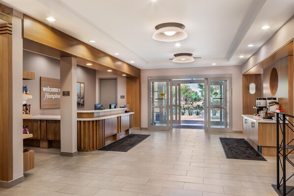 A hotel reception area with a modern design, featuring a reception desk, a 'Welcome to Hampton' sign, and a view of the outdoor area through glass doors.