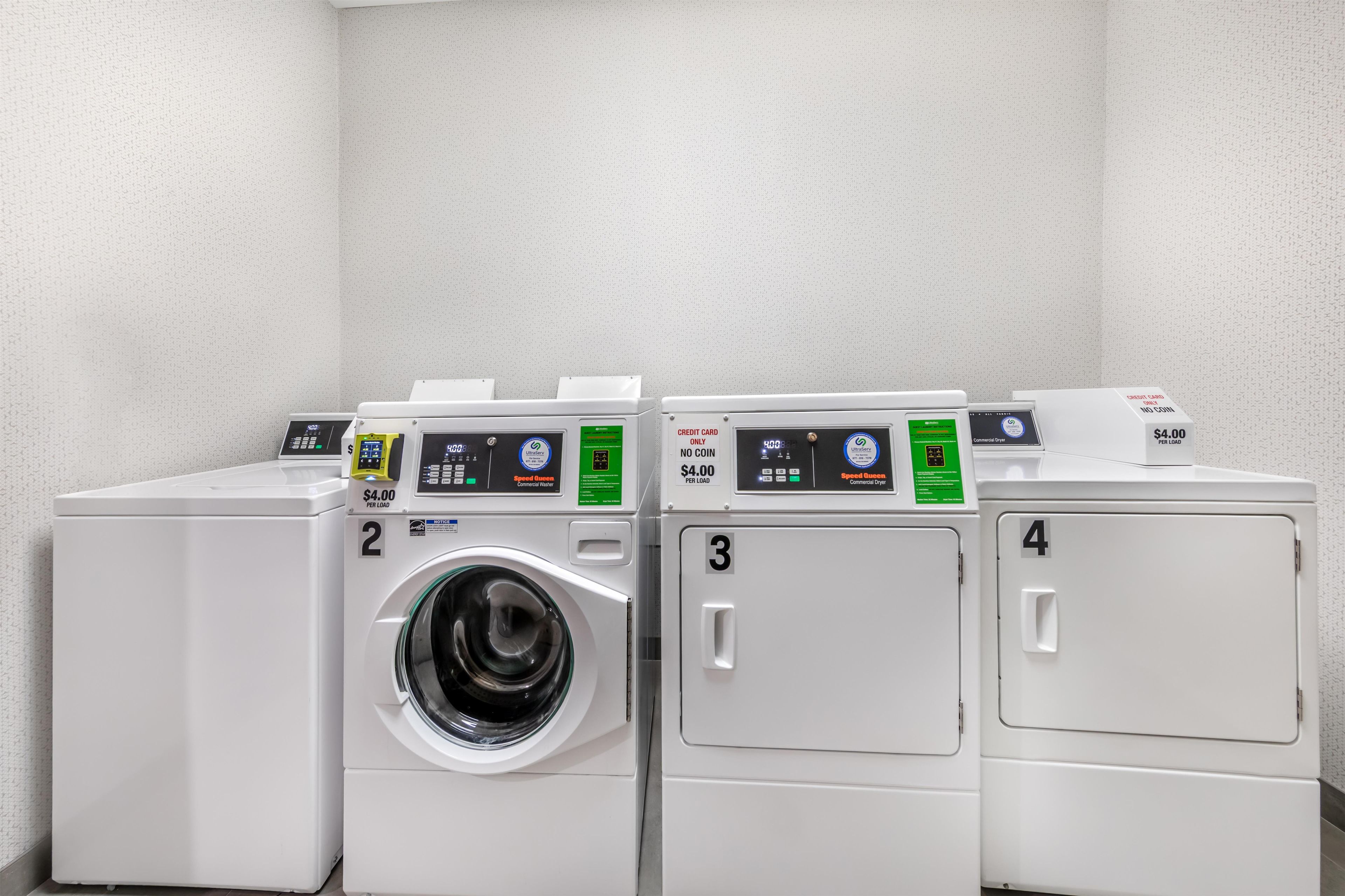 Cuatro lavadoras blancas en fila, con pantallas digitales y puertas numeradas, que señalan un lavadero automático.