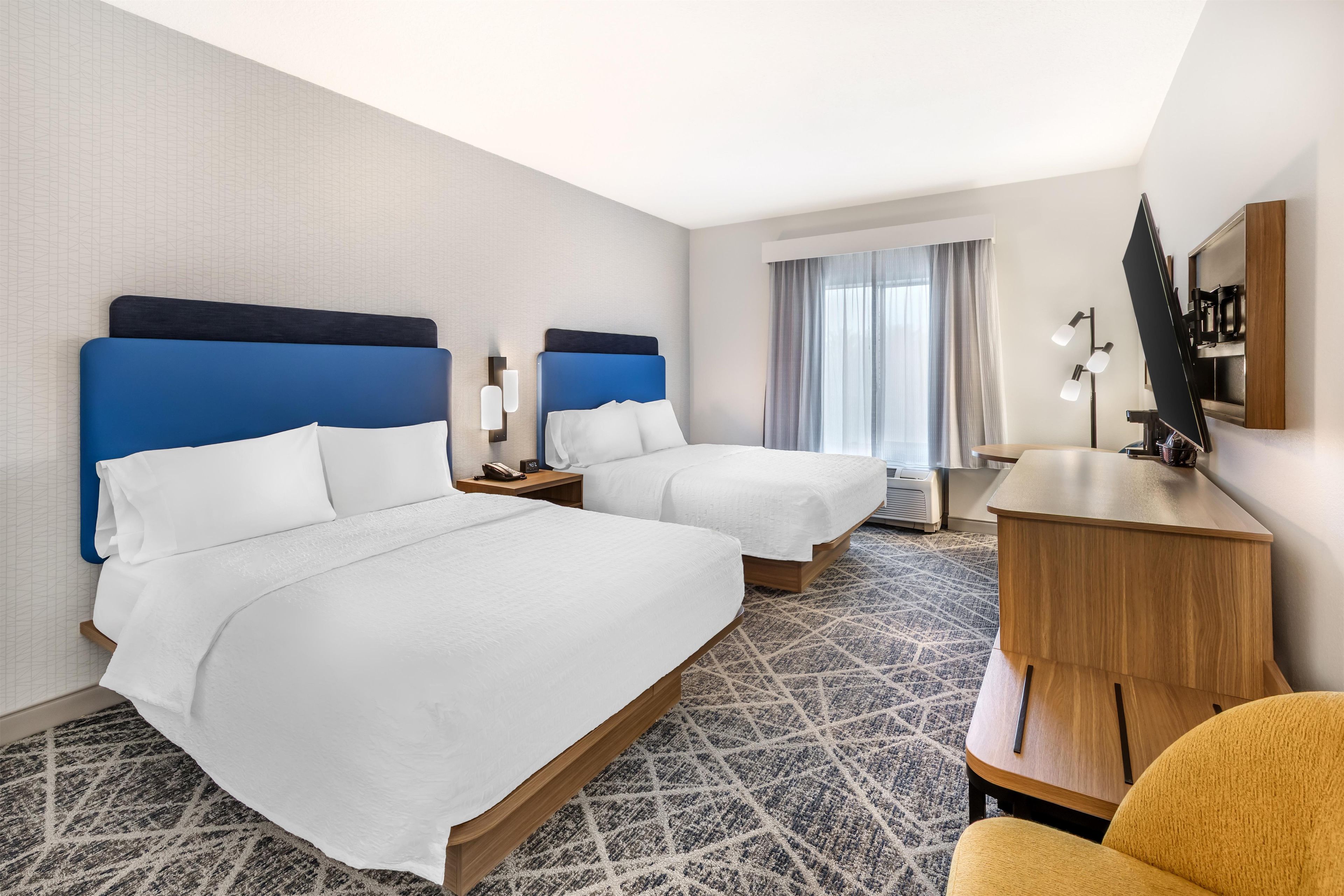 Une chambre d’hôtel avec deux lits, un bureau, une chaise, une télévision et une fenêtre avec des rideaux.